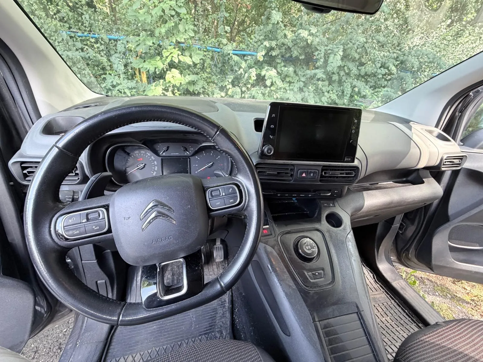 Wnętrze samochodu marki Citroën widziane z perspektywy kierowcy. Widoczna jest czarna kierownica z logo marki i napisem AIRBAG, deska rozdzielcza z ekranem multimedialnym, zegary analogowe oraz konsola środkowa z pokrętłem. Przez przednią szybę widoczne są zielone liście drzew.