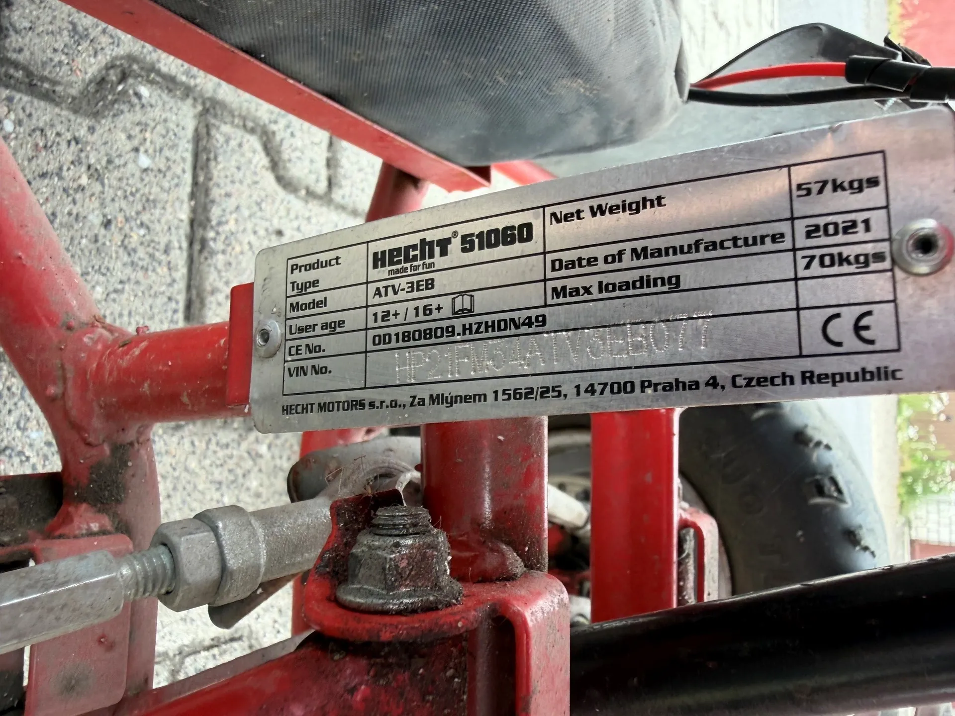 Zbliżenie na metalową tabliczkę znamionową przymocowaną do czerwonej ramy pojazdu. Tabliczka zawiera następujące informacje: Product HECHT 51060, Type made for fun, Model ATV-3EB, User age 12+ / 16+, CE No. 00180809.HZDHN49, VIN No. (numer częściowo zamazany), Net Weight 57kgs, Date of Manufacture 2021, Max loading 70kgs oraz znak CE i adres producenta: HECHT MOTORS s.r.o., Za Mýgnem 1562125, 14700 Praha 4, Czech Republic. Tabliczka jest zorientowana pionowo, ale tekst na niej jest obrócony o 90 stopni w lewo.