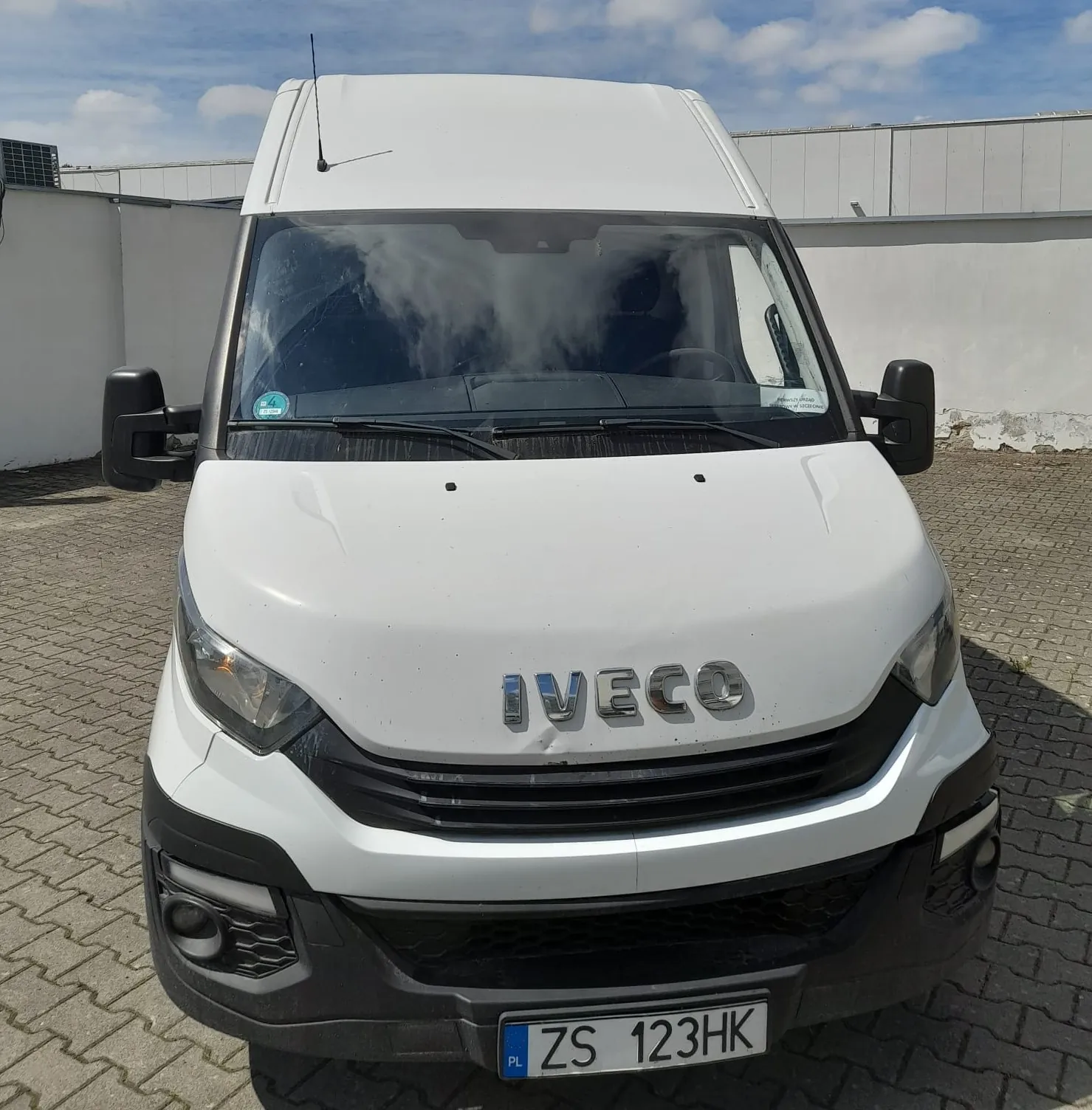 Widok z przodu białego samochodu dostawczego marki Iveco. Na masce widnieje srebrny napis 'IVECO'. Pojazd posiada czarny zderzak oraz tablicę rejestracyjną o numerze 'ZS 123HK'. Samochód stoi na kostce brukowej na tle białego budynku pod lekko zachmurzonym niebem.