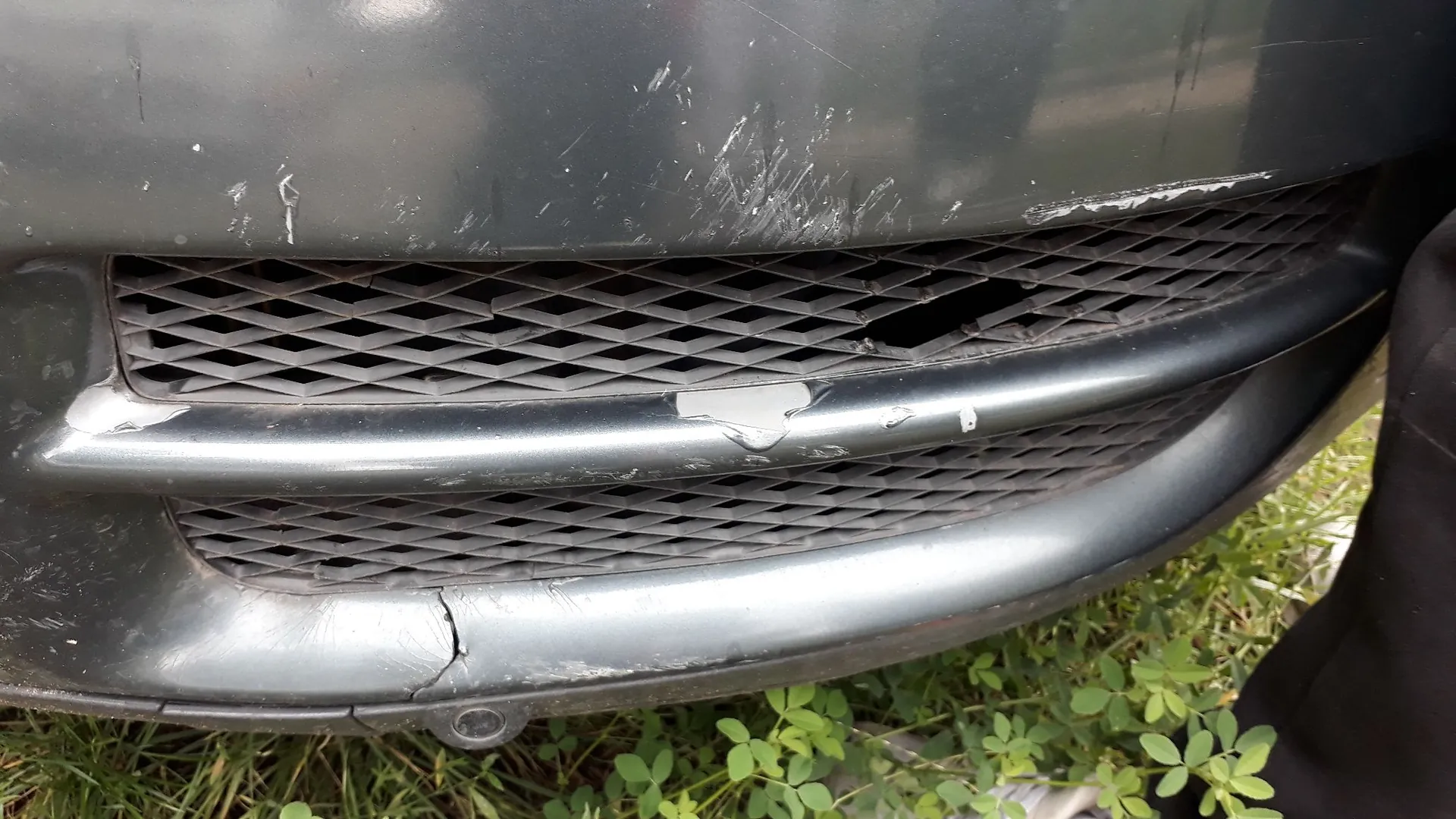 Zbliżenie na uszkodzony przedni zderzak ciemnoszarego samochodu. Na lakierze widoczne są liczne zarysowania i odpryski farby. Plastikowa kratka wlotu powietrza o strukturze plastra miodu jest pęknięta i posiada dziurę w górnej części. Dolna krawędź zderzaka jest porysowana i lekko odkształcona. W tle widoczna jest zielona trawa.