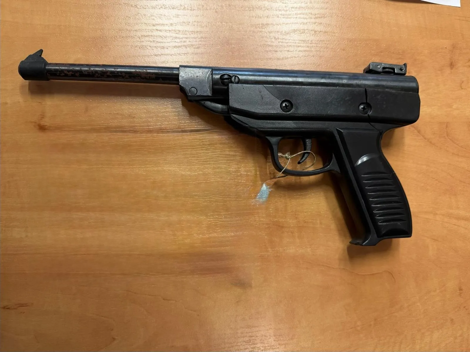 Czarny pistolet leżący poziomo na drewnianym blacie. Broń posiada długą lufę z charakterystycznym zakończeniem, metalową obudowę zamka oraz plastikową rękojeść z żłobieniami. Do spustu przywiązana jest mała, biała etykieta na sznurku.
