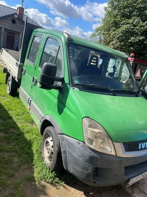Zielony samochód dostawczy typu furgon z otwartą platformą ładunkową z tyłu, zaparkowany na trawiastym poboczu. Widoczny jest przód pojazdu z dużymi reflektorami, czarnym zderzakiem oraz fragmentem tablicy rejestracyjnej z tekstem 'E3P PN8'. W tle widać fragment budynku oraz zielone drzewa pod niebem z białymi chmurami.
