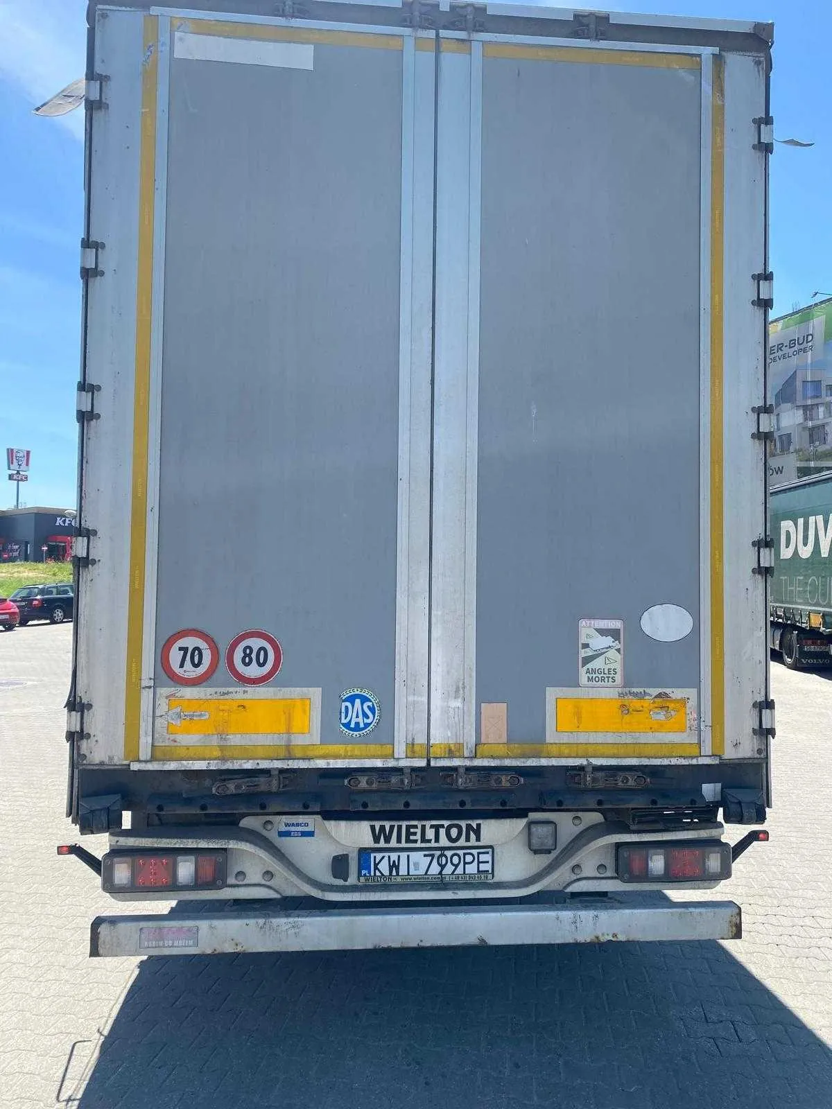 Tylny widok szarej naczepy ciężarówki marki Wielton. Na drzwiach naczepy znajdują się okrągłe znaki ograniczenia prędkości: 70 i 80 km/h, niebieska naklejka z napisem 'DAS' oraz znak ostrzegawczy 'ANGLES MORTS'. Na dole znajduje się tablica rejestracyjna o numerze KWI 799PE oraz napis 'WIELTON'.