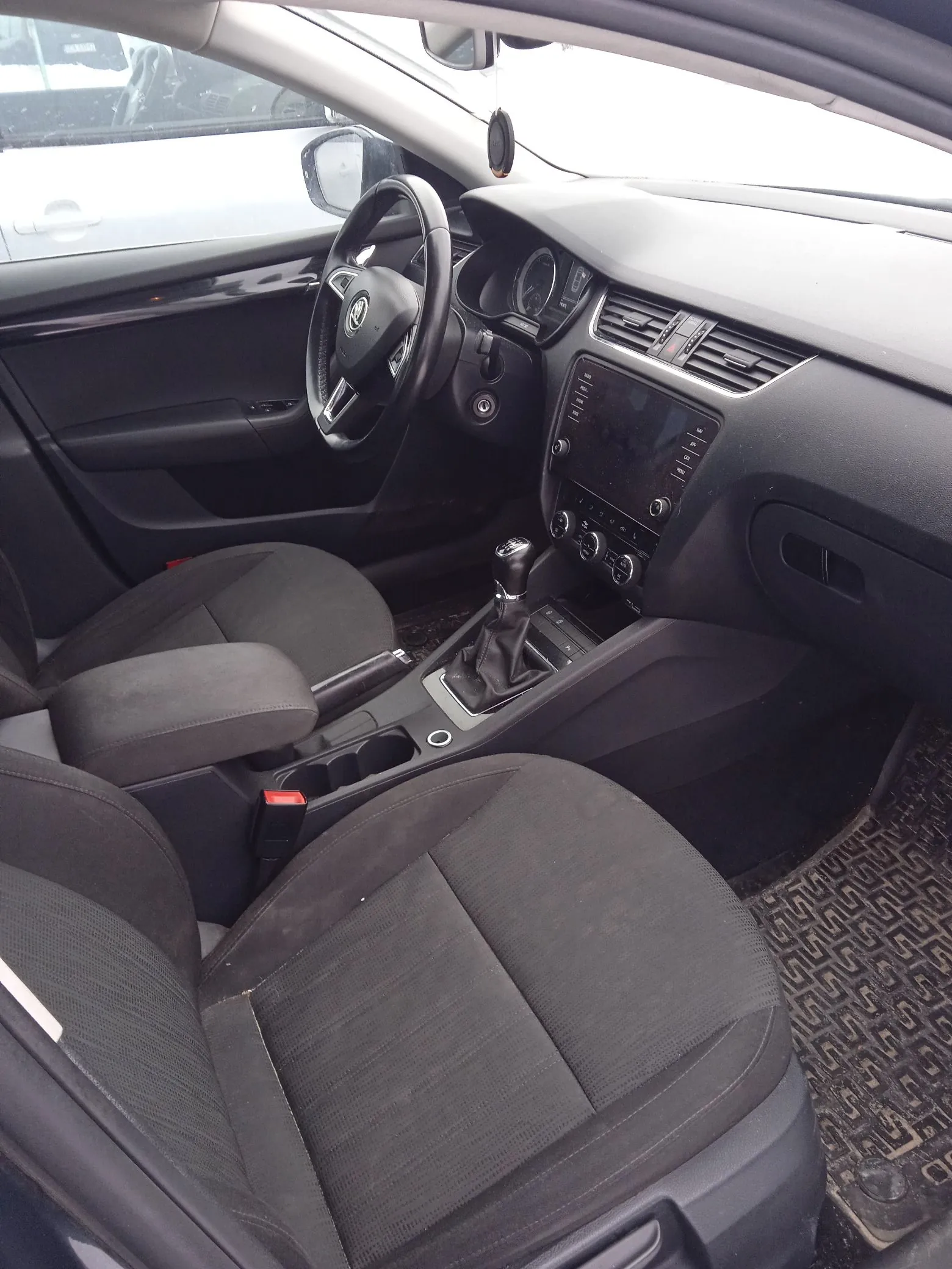 Wnętrze samochodu marki Skoda, widok z perspektywy pasażera na przedni rządek. Widoczna jest czarna kierownica z logo marki, deska rozdzielcza z ekranem multimedialnym, konsola środkowa z dźwignią zmiany biegów oraz dwa ciemnoszare, materiałowe fotele. Na podłodze znajduje się gumowa mata z wzorem.