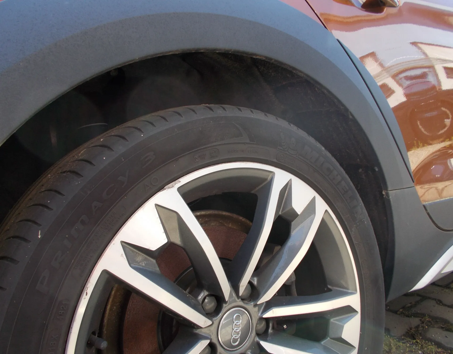 Zbliżenie na koło samochodu z oponą marki Michelin model Primacy 3 oraz srebrną felgą aluminiową z logo Audi w centrum. Koło znajduje się w nadkolu brązowego samochodu z ciemnoszarą plastikową obudową nadkola.