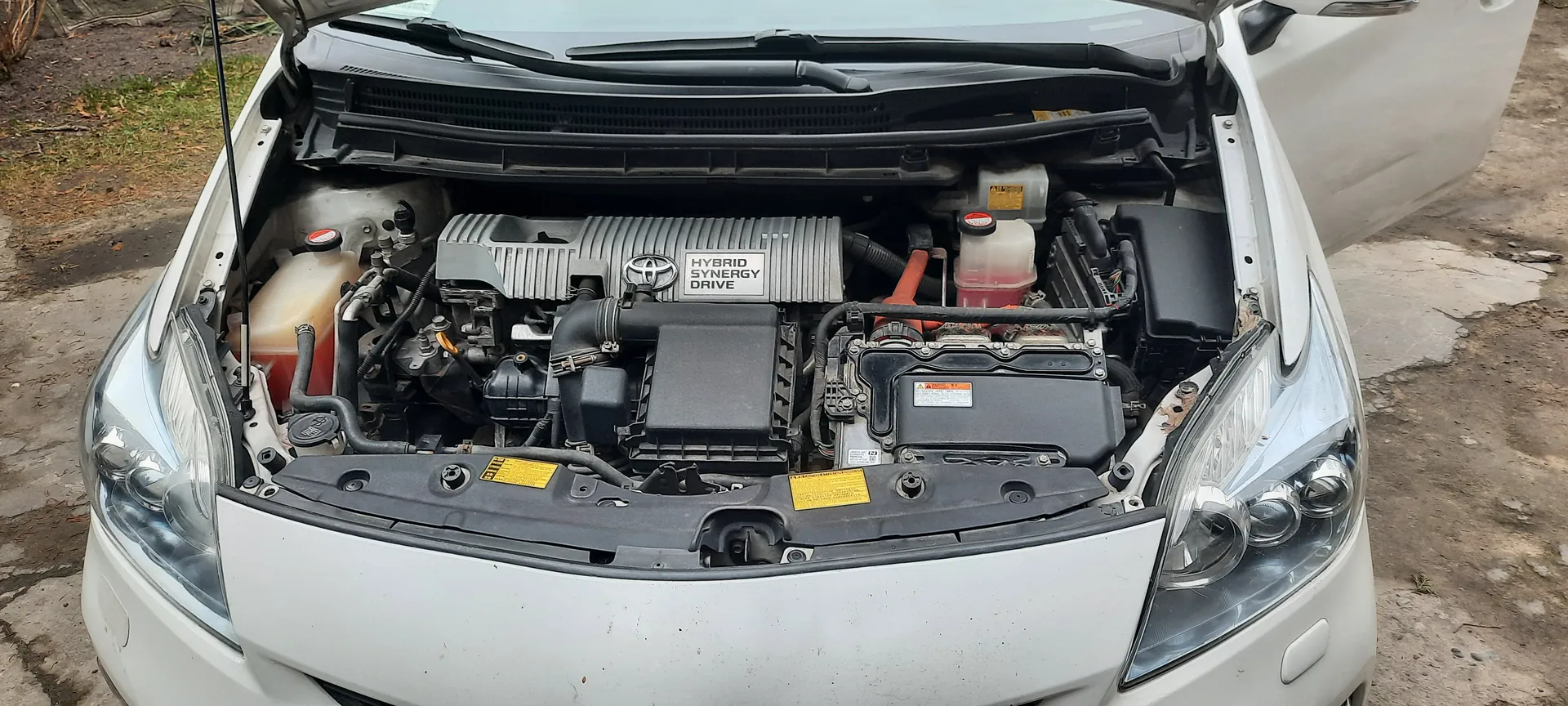 Biały samochód z otwartą maską przedstawiający silnik hybrydowy. W centralnej części silnika widoczny napis 'HYBRID SYNERGY DRIVE' oraz logo Toyoty. W silniku znajdują się zbiorniki z czerwonymi korkami, czarne elementy mechaniczne oraz żółte etykiety ostrzegawcze. Tło stanowi nierówna powierzchnia ziemi z fragmentami trawy i betonu.
