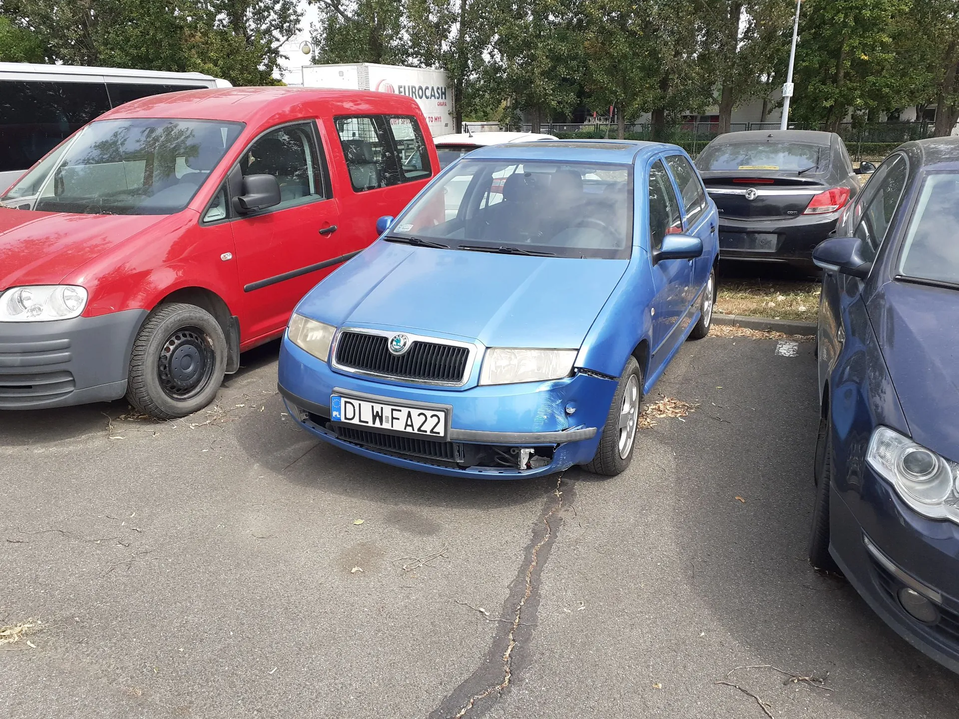 Parking z zaparkowanymi samochodami. Na pierwszym planie znajduje się niebieski samochód marki Skoda z tablicą rejestracyjną DLW FA22, który ma uszkodzony przedni zderzak po prawej stronie. Po lewej stronie stoi czerwony samochód dostawczy, a po prawej ciemnoniebieski samochód. W tle widoczny jest biały samochód oraz biała naczepa z napisem EUROCASH.
