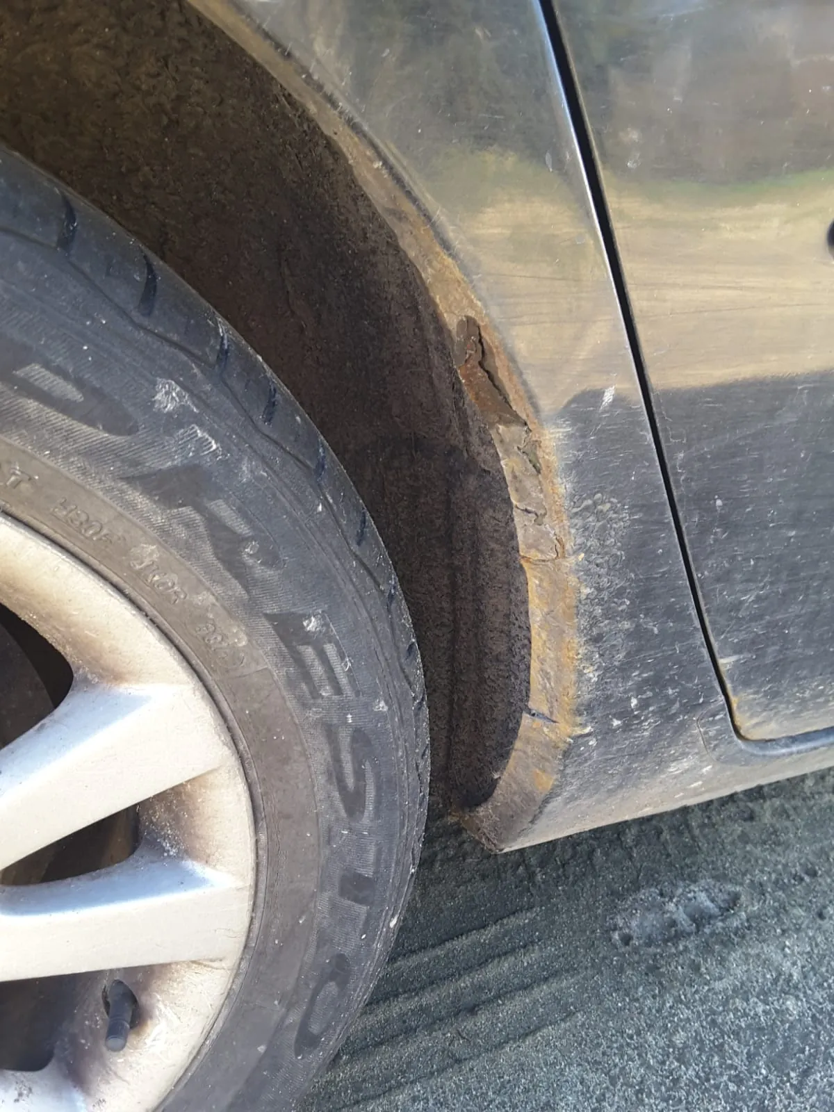 Zbliżenie na koło samochodu i nadkole. Opona pokazuje widoczny zużycie z częściowym napisem "EST". Nadkole ma znaczne ślady rdzy i uszkodzeń, w tym odłupanej farby i zardzewiałego metalu. Karoseria samochodu jest ciemna i zabrudzona. Podłoga to asfalt z widocznymi śladami opon.
