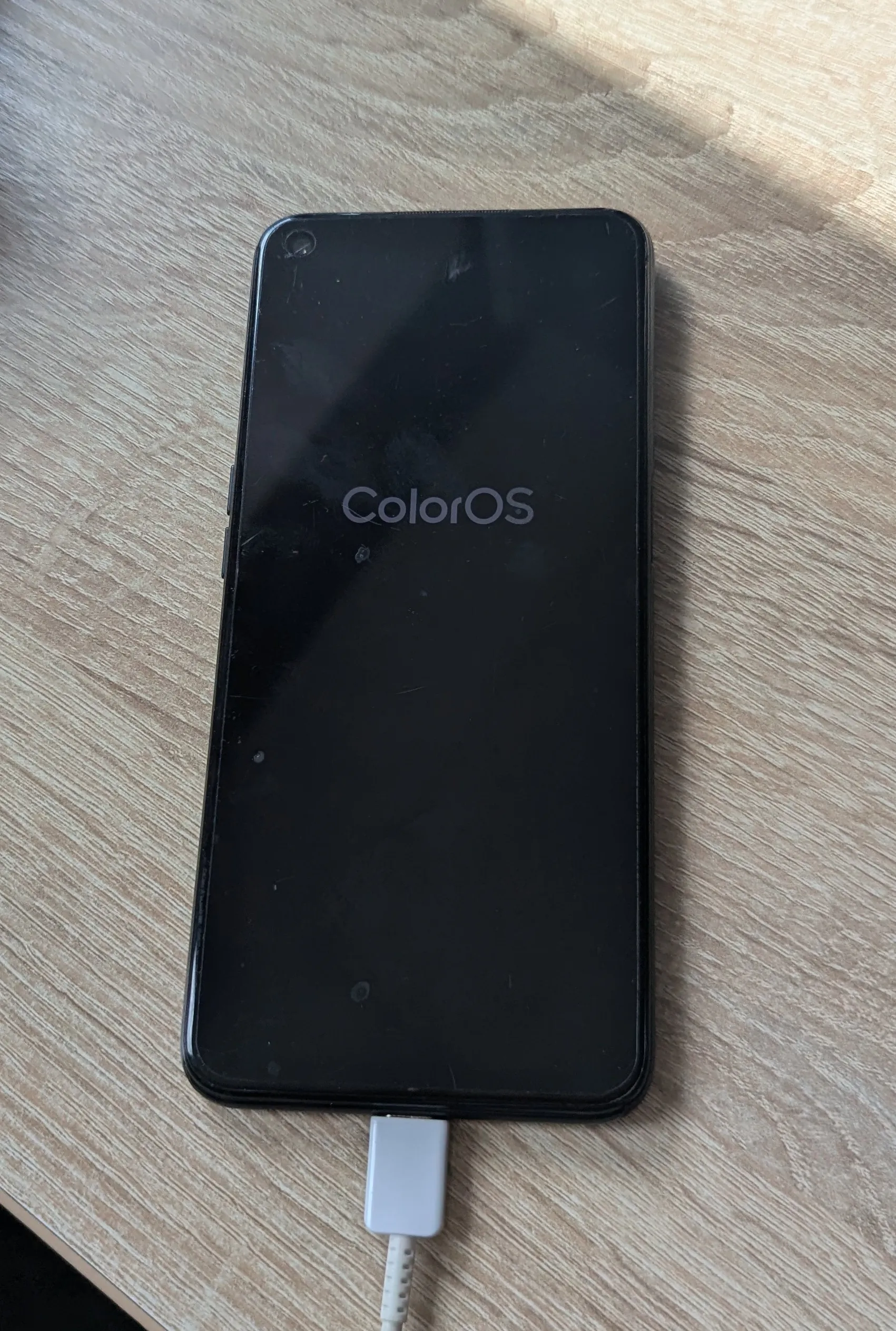 Czarny smartfon leżący na drewnianym blacie. Na ekranie urządzenia widnieje biały napis „ColorOS”. Do dolnej krawędzi telefonu podłączony jest biały kabel ładowania USB.