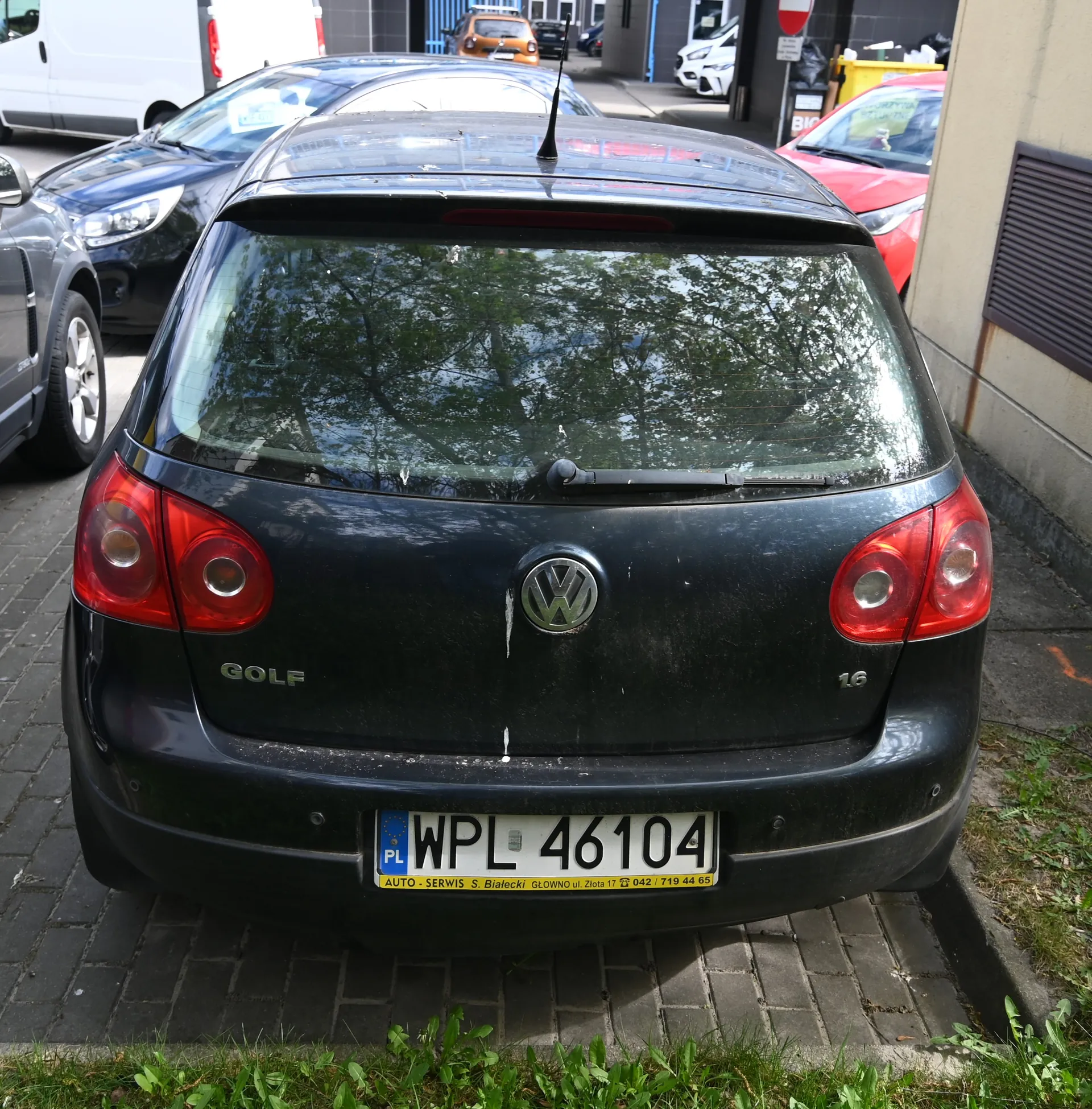 Z tyłu czarny samochód Volkswagen Golf z widocznym logo VW, napisem GOLF po lewej stronie i oznaczeniem 1.6 po prawej. Tablica rejestracyjna PL WPL 46104 z żółtym pasem poniżej zawierającym tekst: AUTO - SERWIS S. Białecki GŁOWNO ul. Złota 17 042 / 719 44 65. W tle zaparkowane samochody (m.in. ciemnoniebieski, czerwony, biały van), budynki, żółty pojemnik na śmieci, znak zakazu wjazdu oraz napis BIO na tablicy. Samochód stoi na brukowanej powierzchni, u dołu widoczna trawa.