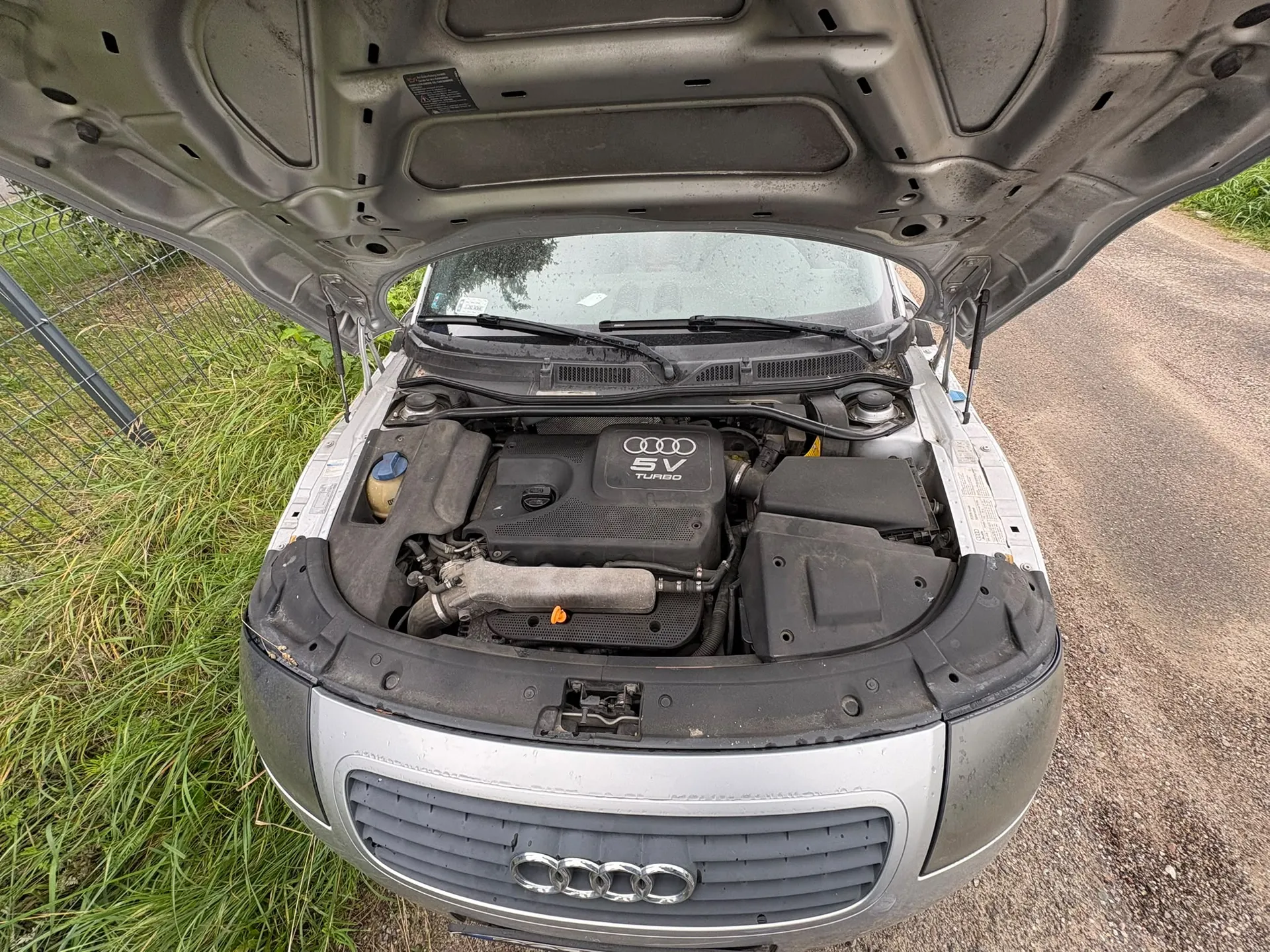 Widok z przodu na otwartą maskę srebrnego samochodu marki Audi. Wewnątrz komory silnika widoczna jest czarna osłona silnika z logo Audi oraz napisem '5V TURBO'. Samochód stoi na piaszczystej drodze obok zielonej trawy i metalowego ogrodzenia.
