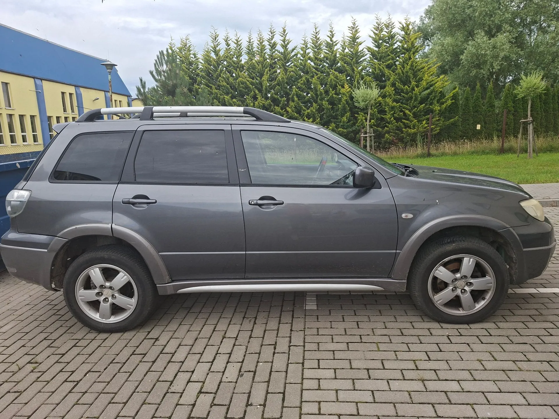 Szary samochód typu SUV zaparkowany bokiem na kostce brukowej. Pojazd posiada srebrne relingi dachowe oraz srebrne listwy u dołu nadwozia. W tle widoczny jest fragment niebiesko-żółtego budynku oraz rząd wysokich, zielonych drzew iglastych pod zachmurzonym niebem.