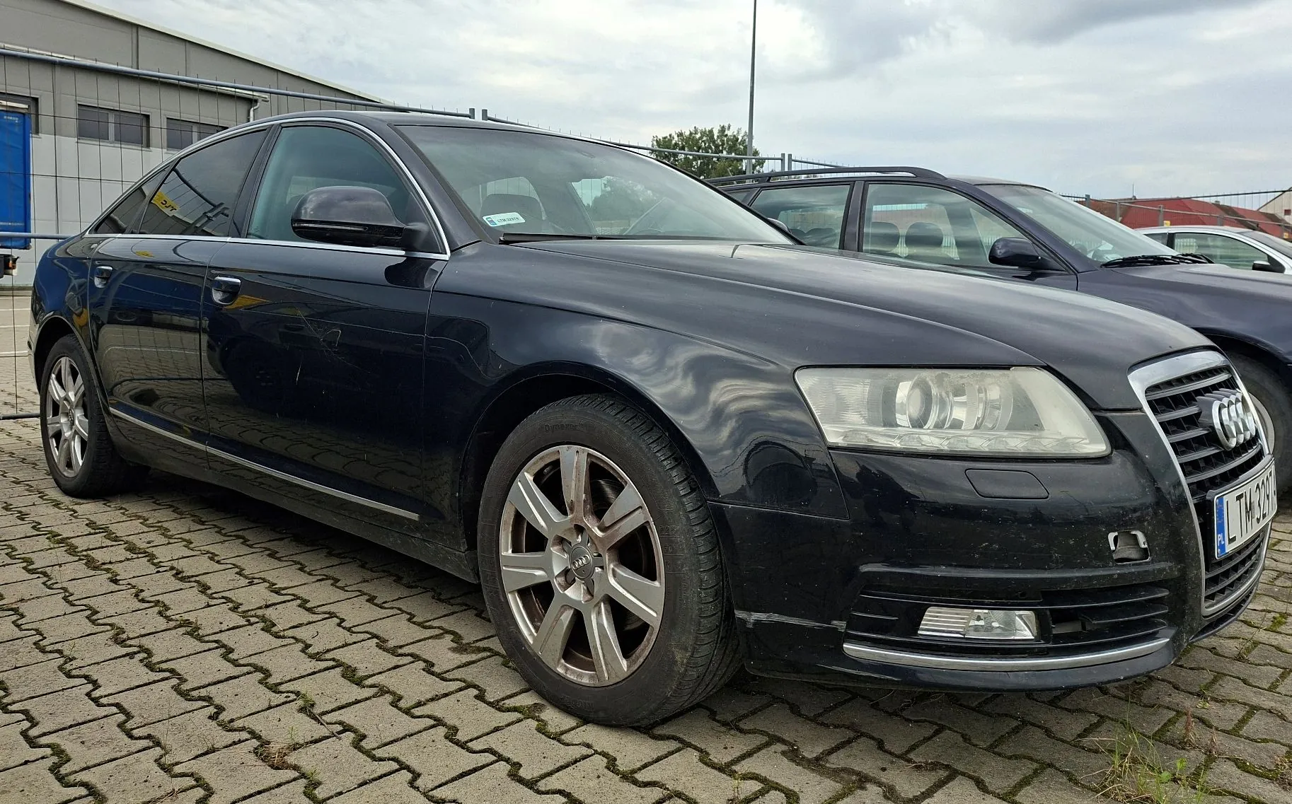 Czarny samochód marki Audi zaparkowany na kostce brukowej przed budynkiem przemysłowym. Pojazd jest widoczny z przodu i z boku, z widoczną tablicą rejestracyjną zaczynającą się od liter LT. W tle widoczny jest fragment innego, ciemnego samochodu oraz szary budynek.