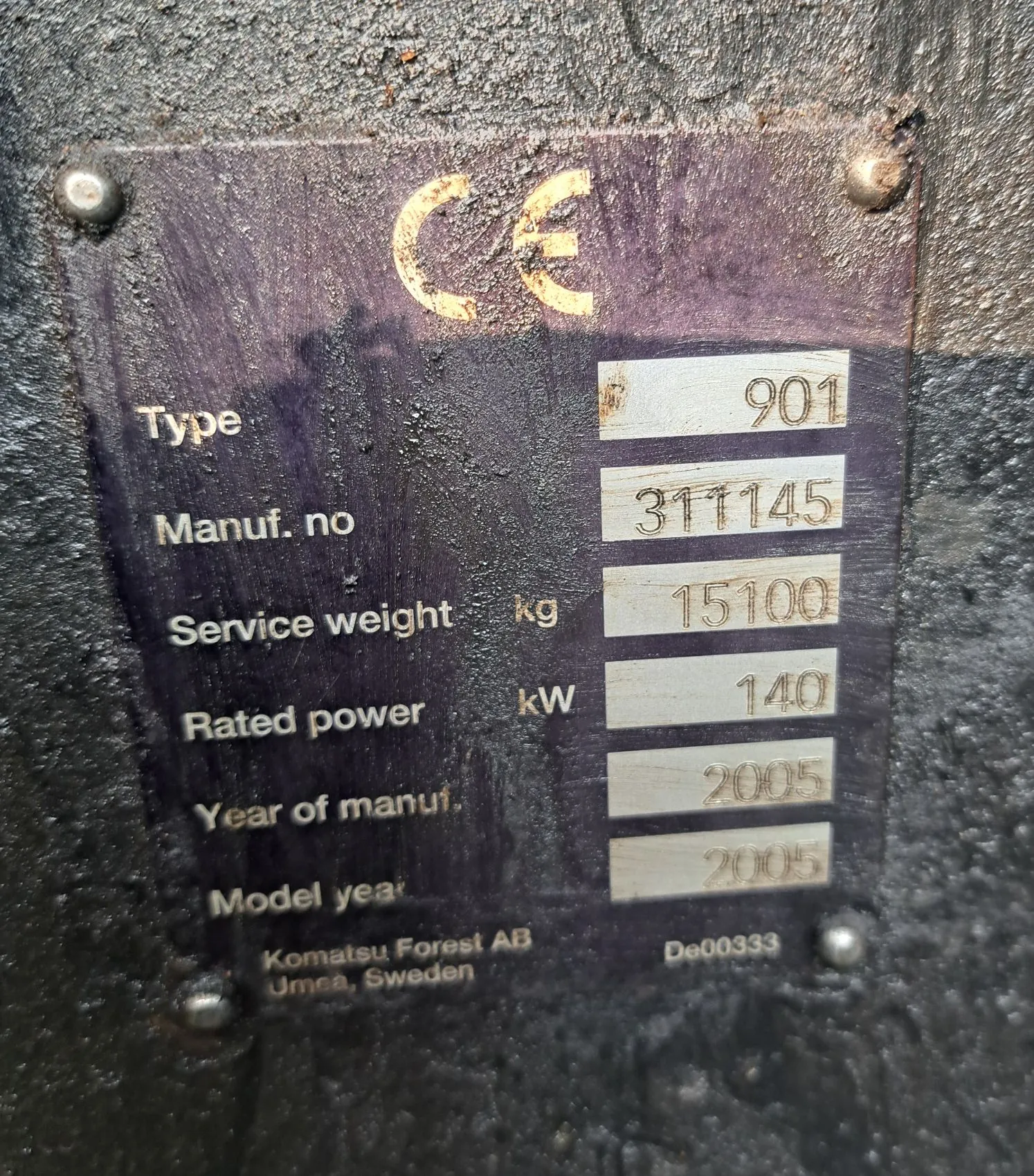 Metalowa tabliczka znamionowa przytwierdzona nitami do ciemnej, chropowatej powierzchni. Na górze znajduje się znak CE. Tabliczka zawiera następujące dane: Type: 901, Manuf. no: 311145, Service weight kg: 15100, Rated power kW: 140, Year of manuf.: 2005, Model year: 2005. Na dole widnieje napis: Komatsu Forest AB Umea, Sweden oraz kod De00333.