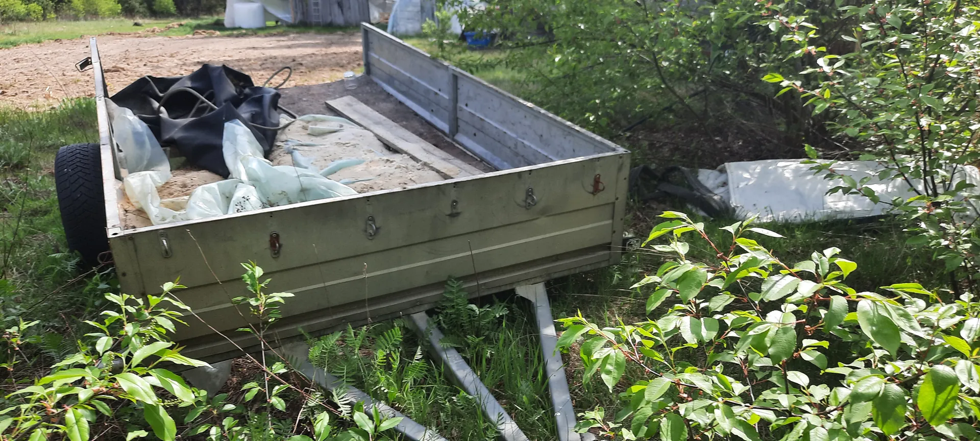 Oliwkowa przyczepa platformowa z metalowymi burtami stojąca na trawie. Wewnątrz przyczepy znajdują się czarne i białe worki z materiału, piasek oraz drewniana deska. Po lewej stronie przyczepy widoczne jest koło. W tle brudna droga, zielone krzewy, białe namioty lub budynki oraz drzewa. Całość otoczona jest roślinnością.