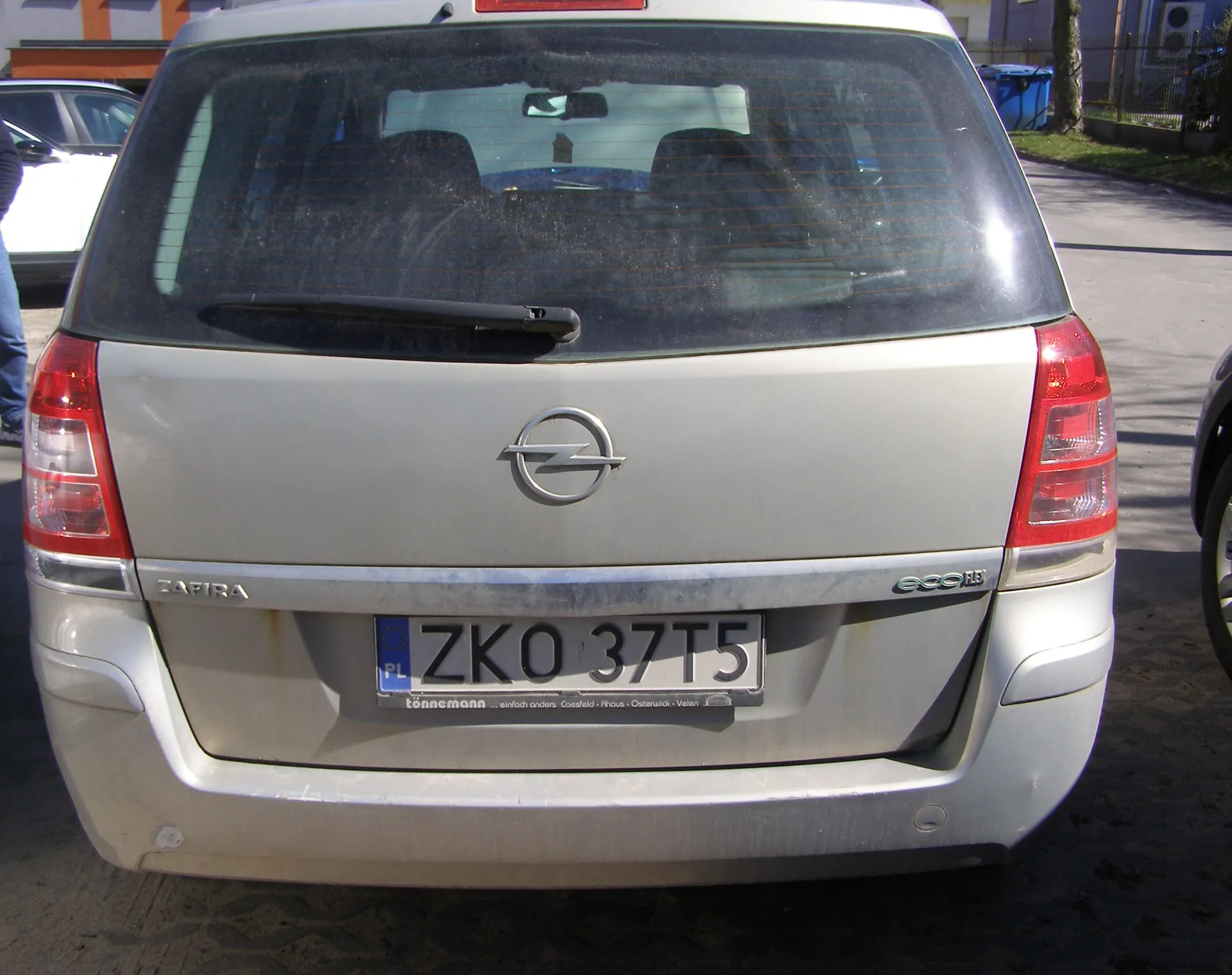 Tył srebrnego samochodu Opel Zafira z widocznym logo marki, napisem ZAFIRA po lewej stronie bagażnika i napisem ecoFLEX po prawej. Tablica rejestracyjna PL ZKO 37T5 z niebieskim pasem po lewej stronie. W tle częściowo widoczne inne pojazdy, budynki i niebieski pojemnik na śmieci. Samochód stoi na asfaltowej powierzchni.
