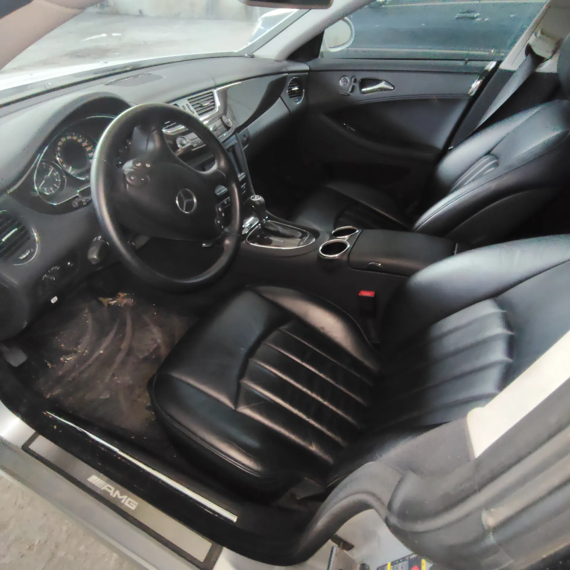 Wnętrze samochodu z widokiem na stronę kierowcy. Widoczna czarna skórzana tapicerka foteli, czarna kierownica z logo Mercedes-Benz, deska rozdzielcza oraz konsola środkowa z uchwytami na kubki. Na progu drzwi widoczny jest napis 'AMG'.