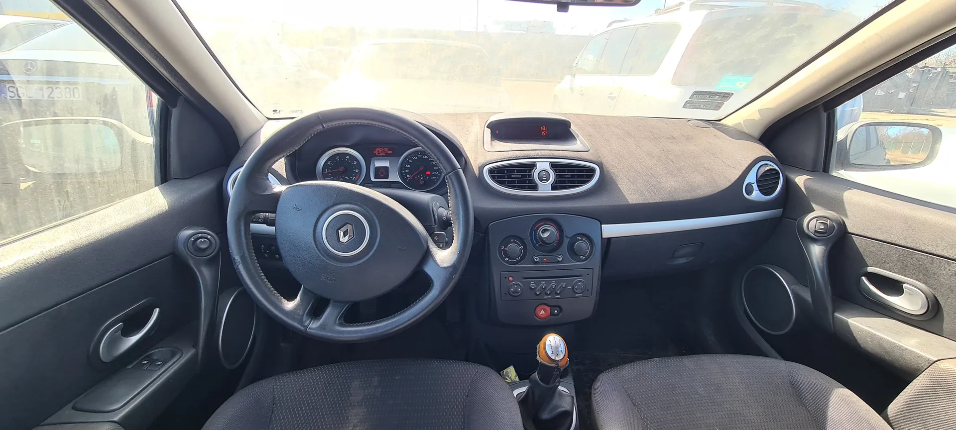 Wnętrze samochodu z widokiem na kierownicę z logo Renault, tablicę rozdzielczą z licznikami prędkości i obrotomierzem, centralną konsolę z przyciskami i gałką zmiany biegów. Widoczne są czarne siedzenia, deska rozdzielcza w kolorze szaro-czarnym oraz zewnętrzne samochody za przednią szybą. Na szybie bocznej widoczny jest fragment tablicy rejestracyjnej SGL 12380. Na wyświetlaczu centralnym czas 13:31 i temperatura 5°C.