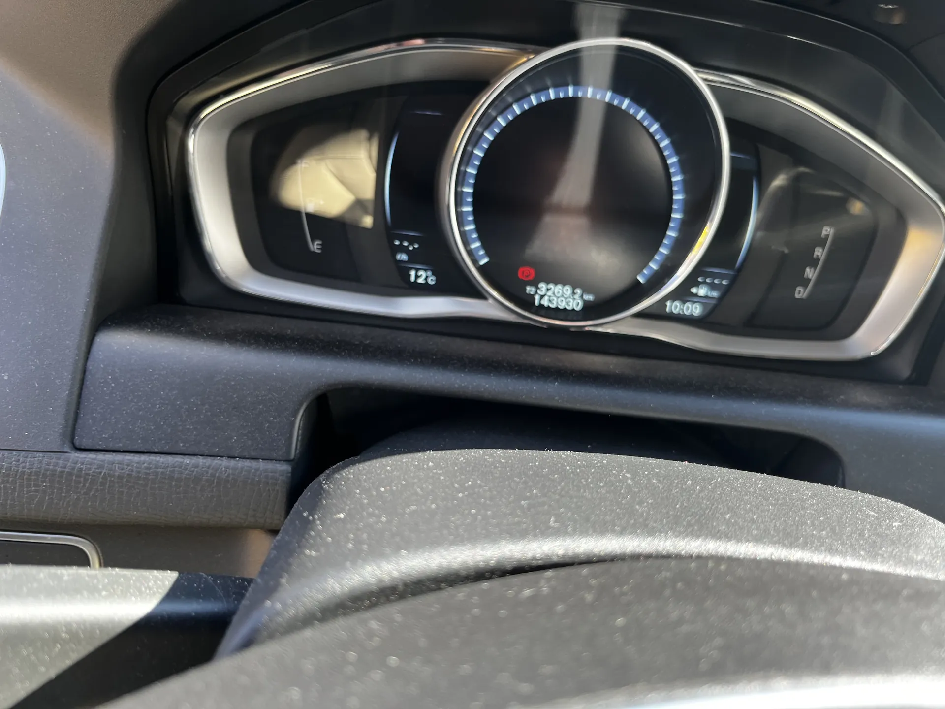 Zbliżenie na deski rozdzielcze samochodu. W centrum znajduje się okrągły wskaźnik prędkości z niebieskim pierścieniem, poniżej widoczne liczby: 3269,2 i 143930. Po lewej stronie wskaźnik paliwa z oznaczeniami E i F, obok temperatura 12°C. Po prawej stronie wskaźnik bieżącego czasu 10:09 oraz oznaczenia biegów P, R, N, D. Na dole widoczna część kierownicy. Całość w kolorze czarnym z srebrnymi elementami dekoracyjnymi.