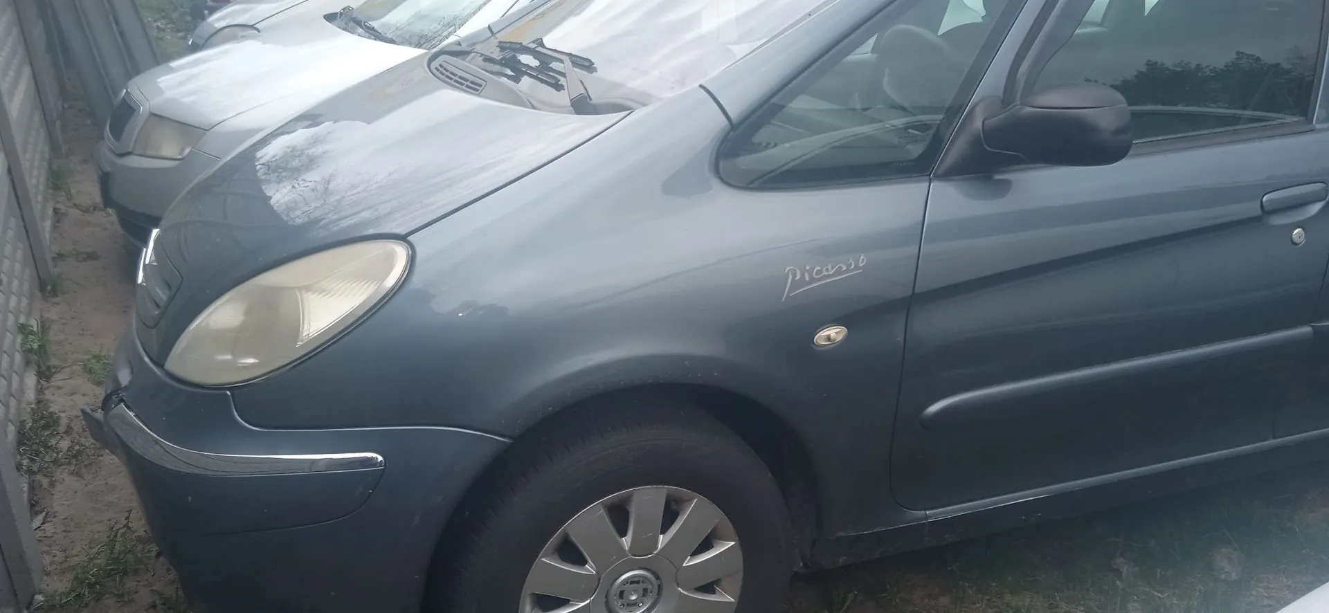 Zbliżenie na przód i bok szarego samochodu marki Citroën model Picasso. Widoczny jest lewy reflektor, przednie koło z felgą aluminiową oraz napis 'Picasso' na przednim błotniku. W tle widoczny jest fragment innego, jasnego samochodu.