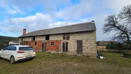 Zdjęcie starego, parterowego budynku gospodarczego o konstrukcji mieszanej z cegły i betonowych bloczków, z dwuspadowym dachem pokrytym eternitem. W budynku znajdują się drewniane drzwi i otwory okienne bez szyb. Przed budynkiem, po lewej stronie, zaparkowany jest biały samochód osobowy typu kombi. W tle widać bezlistne drzewo, trawę oraz niebo z białymi chmurami.
