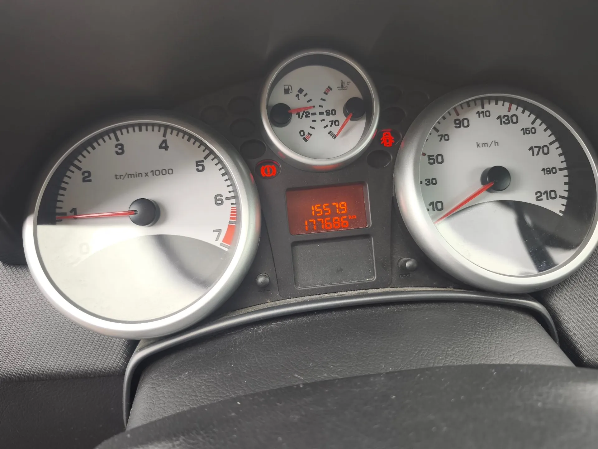 Zbliżenie na deskę rozdzielczą samochodu z trzema okrągłymi wskaźnikami. Po lewej znajduje się obrotomierz z oznaczeniami od 1 do 7 tr/min x 1000. W centrum znajduje się mniejszy wskaźnik poziomu paliwa oraz temperatury silnika, a poniżej czerwony wyświetlacz cyfrowy z liczbami 15579 i 177686. Po prawej stronie znajduje się prędkościomierz z oznaczeniami od 10 do 210 km/h. Na panelu widoczne są czerwone kontrolki ostrzegawcze.