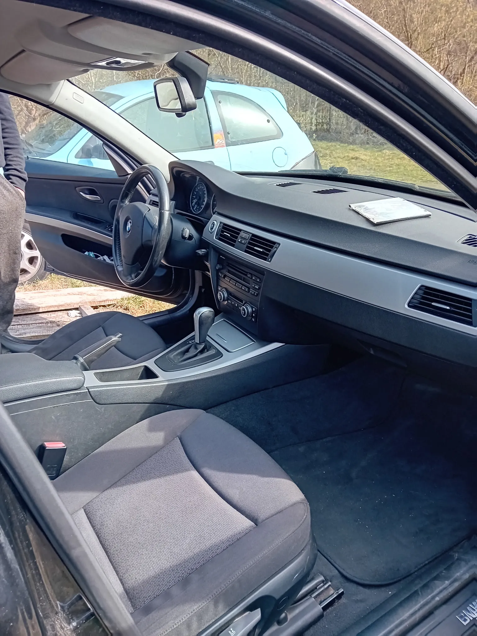 Wnętrze samochodu z widocznym kierownicą z logo BMW, deską rozdzielczą z licznikami, centralną konsolą z przyciskami i dźwignią zmiany biegów. Siedzenia są szare, tapicerowane tkaniną. Drzwi kierowcy są otwarte, za nimi widoczny jest niebieski samochód zaparkowany na trawie oraz suche krzewy. Na desce rozdzielczej leży prostokątny przedmiot. Po lewej stronie kadru widoczna jest część ramienia osoby w ciemnym ubraniu.