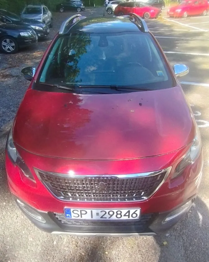 Czerwony samochód osobowy marki Peugeot zaparkowany na parkingu. Widoczny jest przód pojazdu z tablicą rejestracyjną o numerze SPI 29846. W tle znajdują się inne zaparkowane samochody oraz fragmenty drzew.