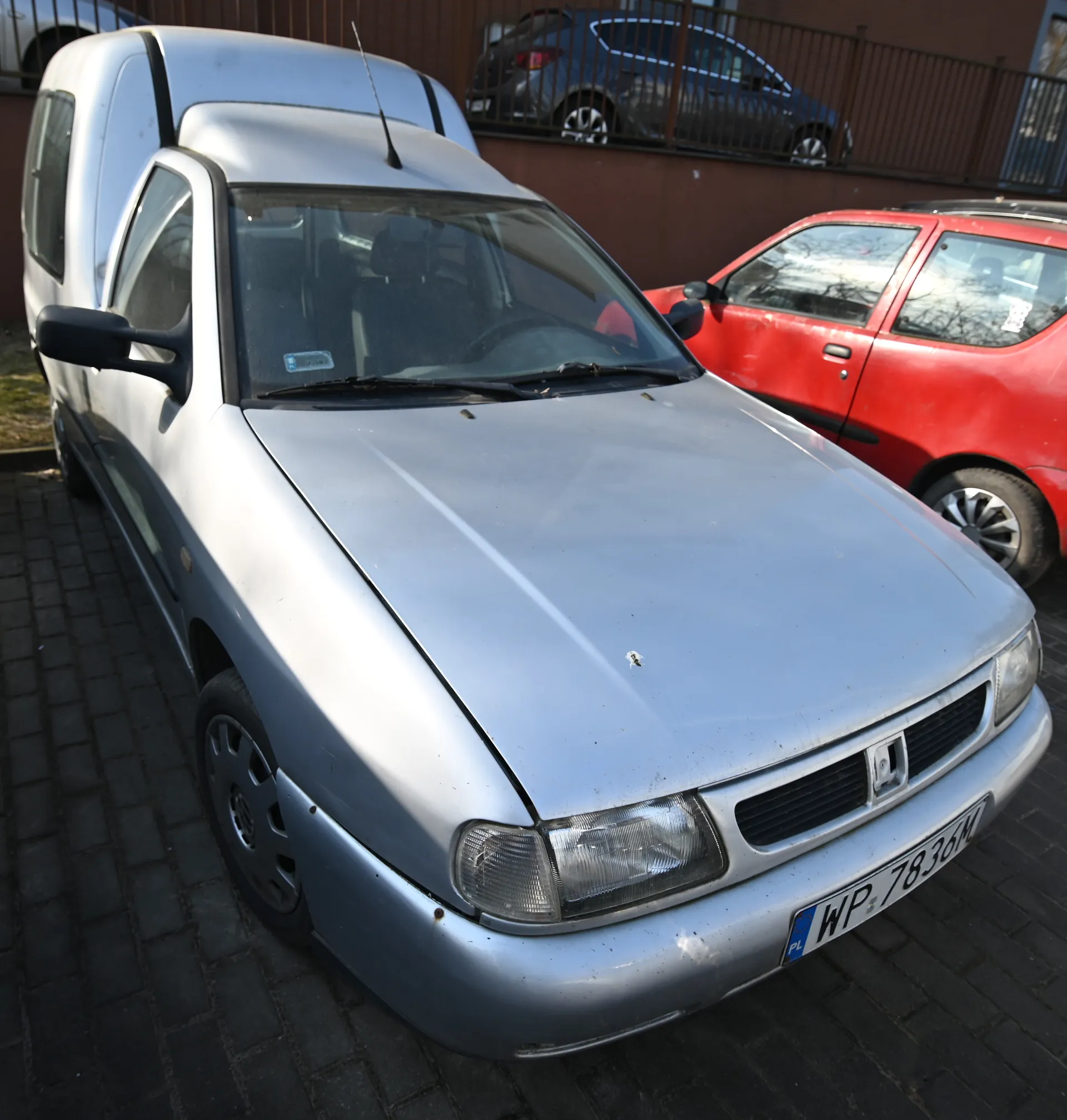 Srebrny samochód osobowy z tablicą rejestracyjną WP 7836M (PL) zaparkowany na brukowanej powierzchni. W tle czerwony samochód, biały furgon oraz ciemnoszary samochód za brązowym ogrodzeniem. Na przedniej szybie srebrnego samochodu widoczny mały naklejony znaczek. Samochody stoją na otwartym obszarze z brązową ścianą i metalowym ogrodzeniem w tle.