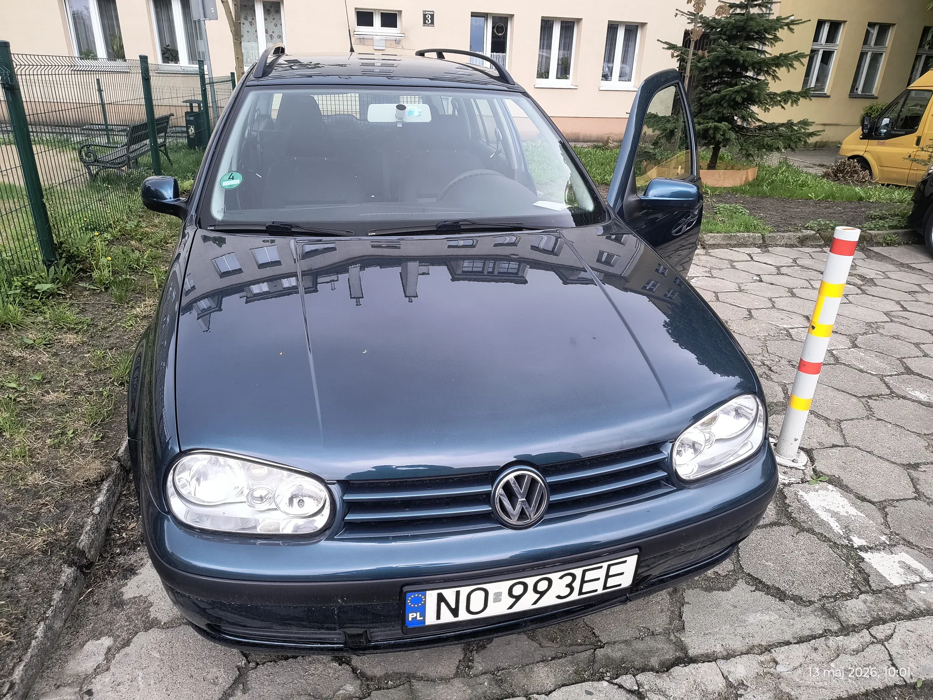 Ciemnoniebieski samochód osobowy marki Volkswagen z otwartą przednią prawą drzwiami, zaparkowany na brukowanej powierzchni. Widoczna tablica rejestracyjna z napisem NO 993EE oraz flagą PL. W tle budynek mieszkalny z oknami, numerem 3, zielone ogrodzenie, ławka, drzewo i żółty samochód dostawczy. Na prawo od pojazdu słup z czerwonymi i żółtymi pasami. W prawym dolnym rogu znacznik daty: 13 maj 2026, 10:01.