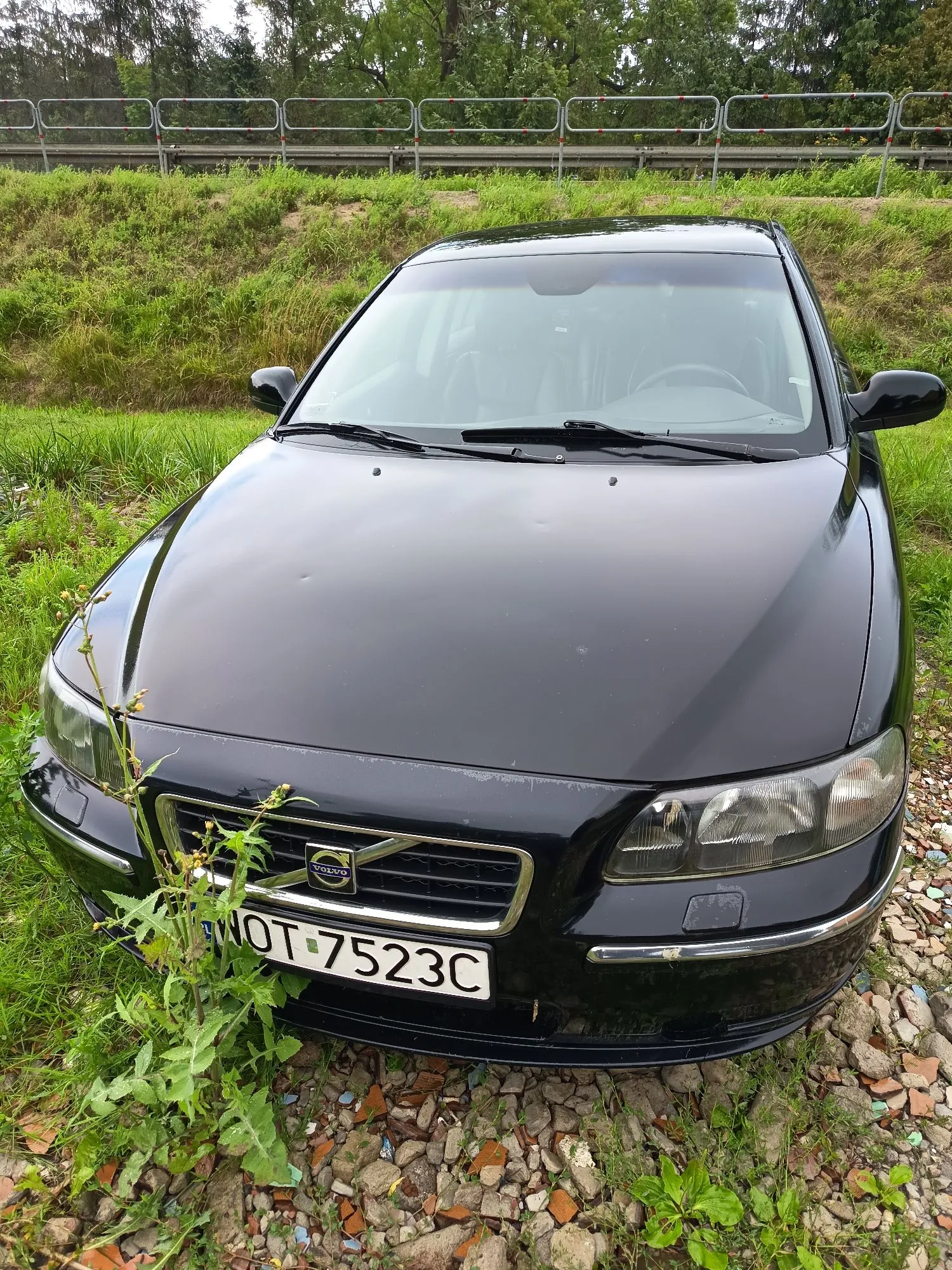 Czarny samochód marki Volvo zaparkowany na trawiastym poboczu obok kamienistej nawierzchni. Widoczny jest przód pojazdu z tablicą rejestracyjną o numerze NOT 7523C. W tle znajduje się zielony nasyp oraz metalowa barierka oddzielająca drogę od terenu zielonego z drzewami.