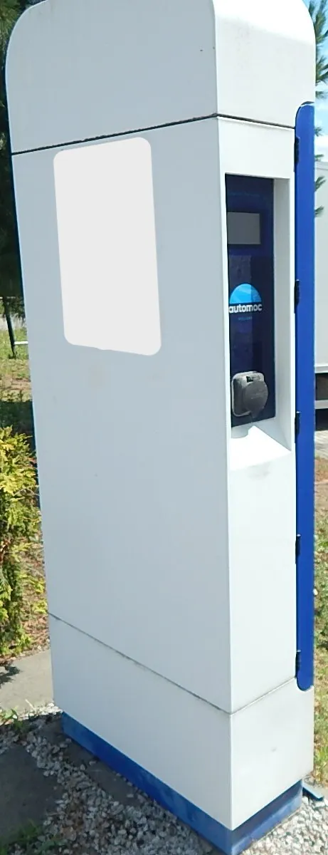 Biały, pionowy automat do ładowania pojazdów elektrycznych. Na urządzeniu znajduje się niebieski panel z napisem 'automoc' oraz gniazdo ładowania. Część przedniej obudowy jest zakryta białym polem. Automat stoi na podłożu z drobnego żwiru.