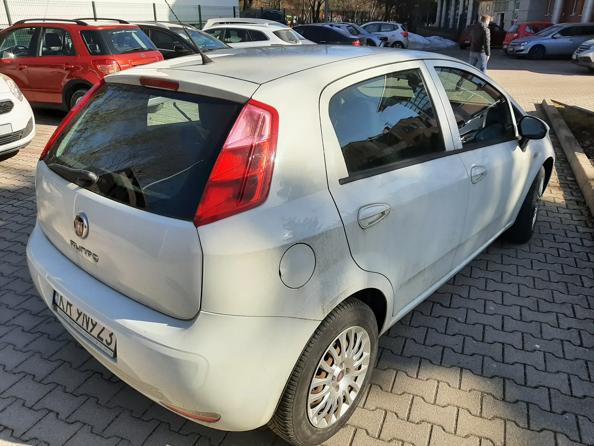 Srebrny samochód Fiat Grande Punto zaparkowany na brukowanym placu parkingowym. Widoczny tył pojazdu z czerwonymi światłami, tablicą rejestracyjną KR 9N9Z3 oraz logo marki. W tle inne zaparkowane samochody (czerwony, biały, czarny), budynki, ogrodzenie i osoba idąca. Słoneczna pogoda, cień na nawierzchni.