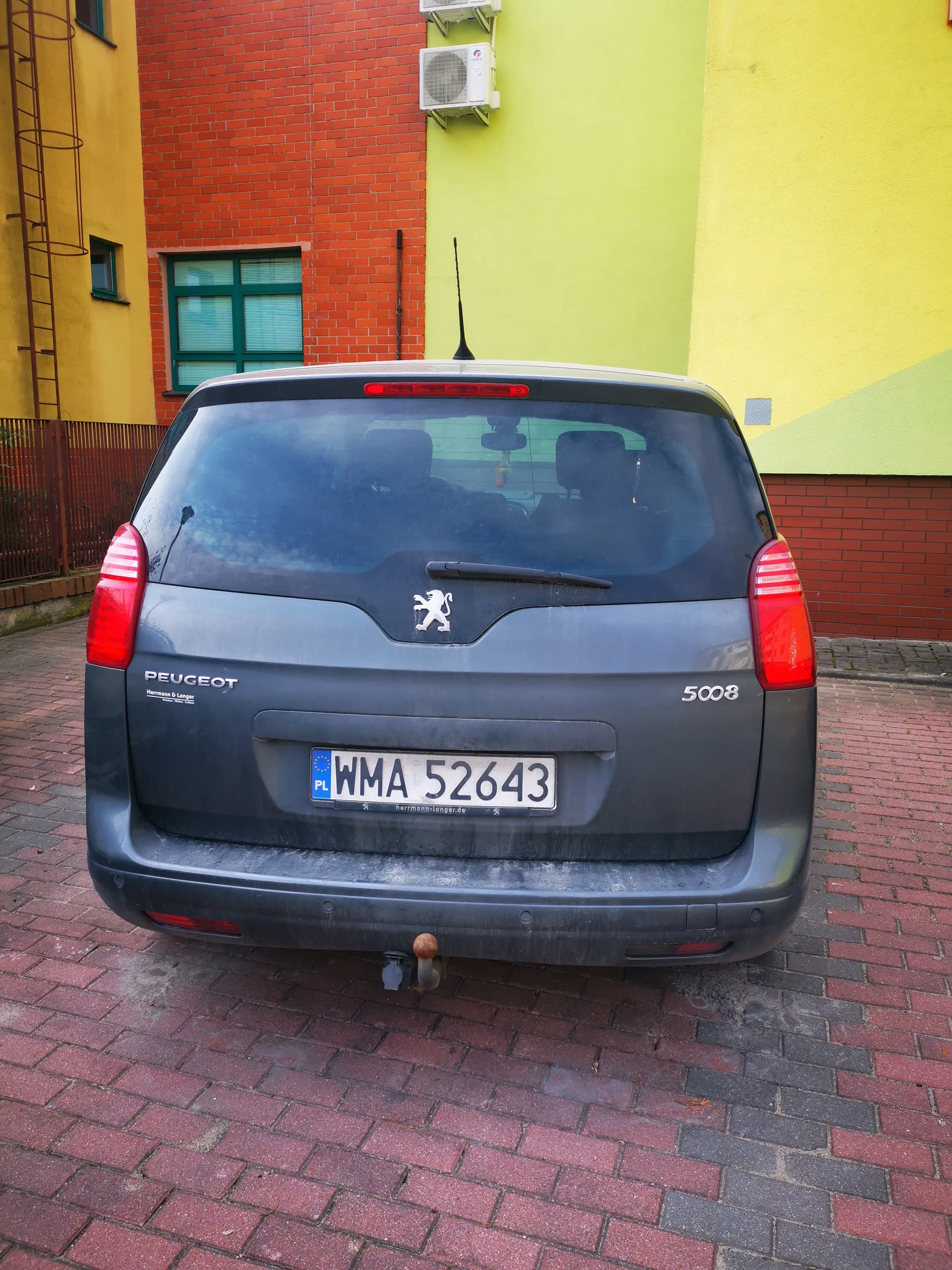 Tył szarego samochodu Peugeot 5008 zaparkowanego na ceglanym placu. Widoczna tablica rejestracyjna PL WMA 52643, logo marki, napisy 'PEUGEOT' i '5008'. W tle budynki z czerwonej cegły, żółtej i zielonej elewacji, okno z zielonymi ramami oraz klimatyzatory. Samochód ma czerwone światła tylna i antenę.