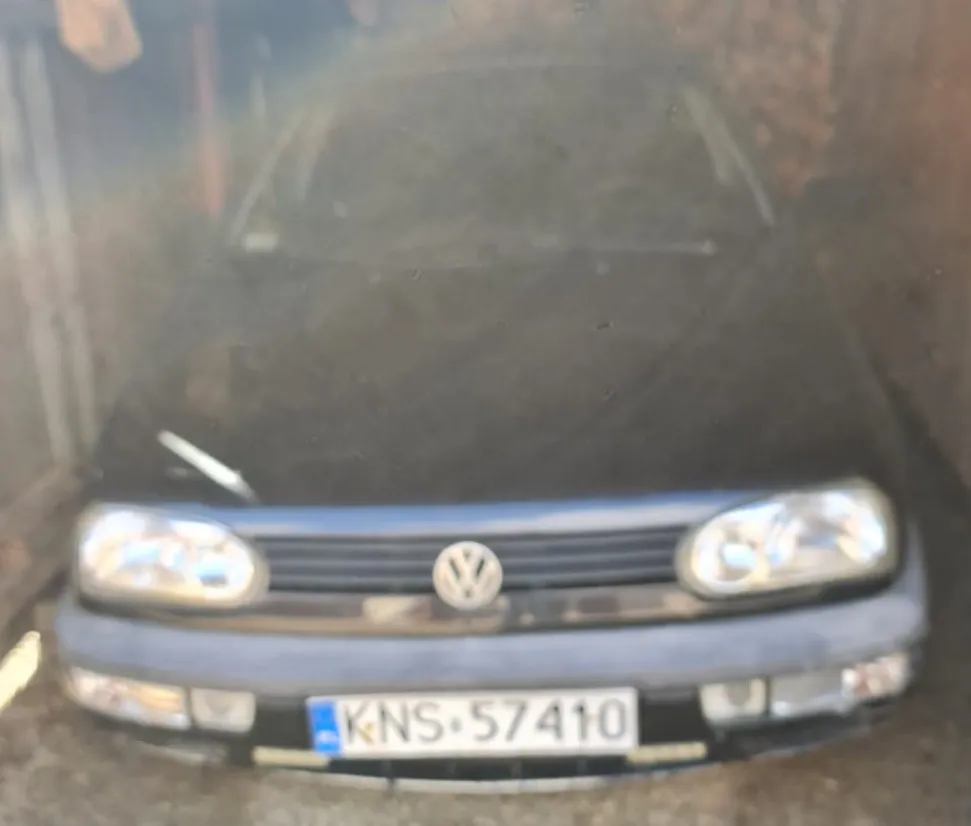 Zdjęcie wykonane przez szybę, przedstawiające przód czarnego samochodu marki Volkswagen. Widoczna jest maska, reflektory oraz tablica rejestracyjna o numerze KNS 57410.