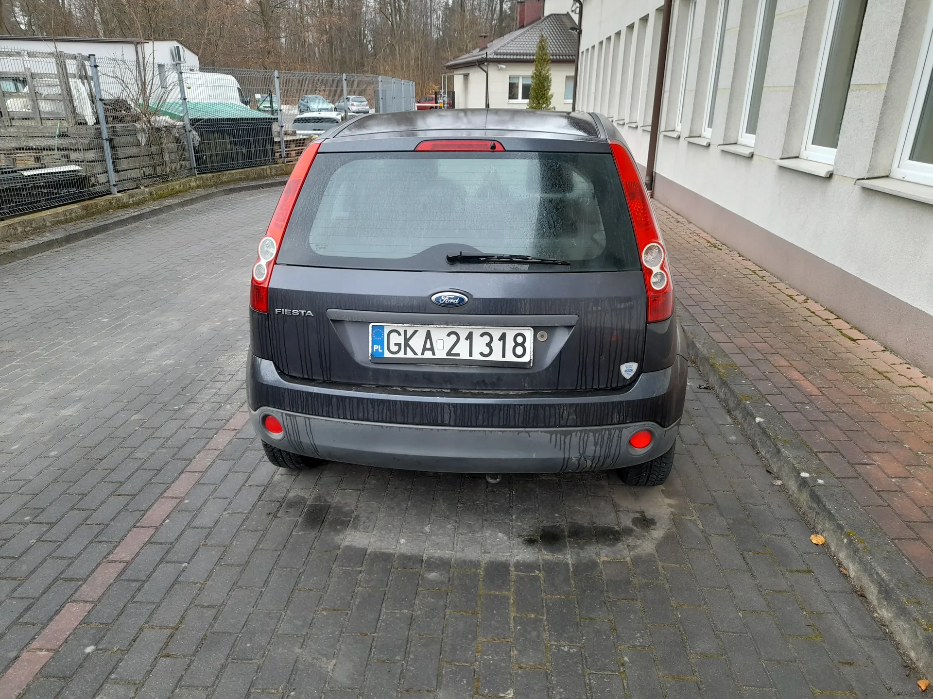 Tył ciemnoszarego samochodu Ford Fiesta zaparkowanego na brukowanej powierzchni. Widoczna tablica rejestracyjna PL GKA 21318, czerwone światła tylnego oświetlenia oraz logo marki. Po prawej stronie budynek z szarymi ścianami i białymi oknami, po lewej metalowe ogrodzenie z innymi pojazdami i drzewami na tle. Na tylnej szybie deszczowa kropla i wycieraczka.