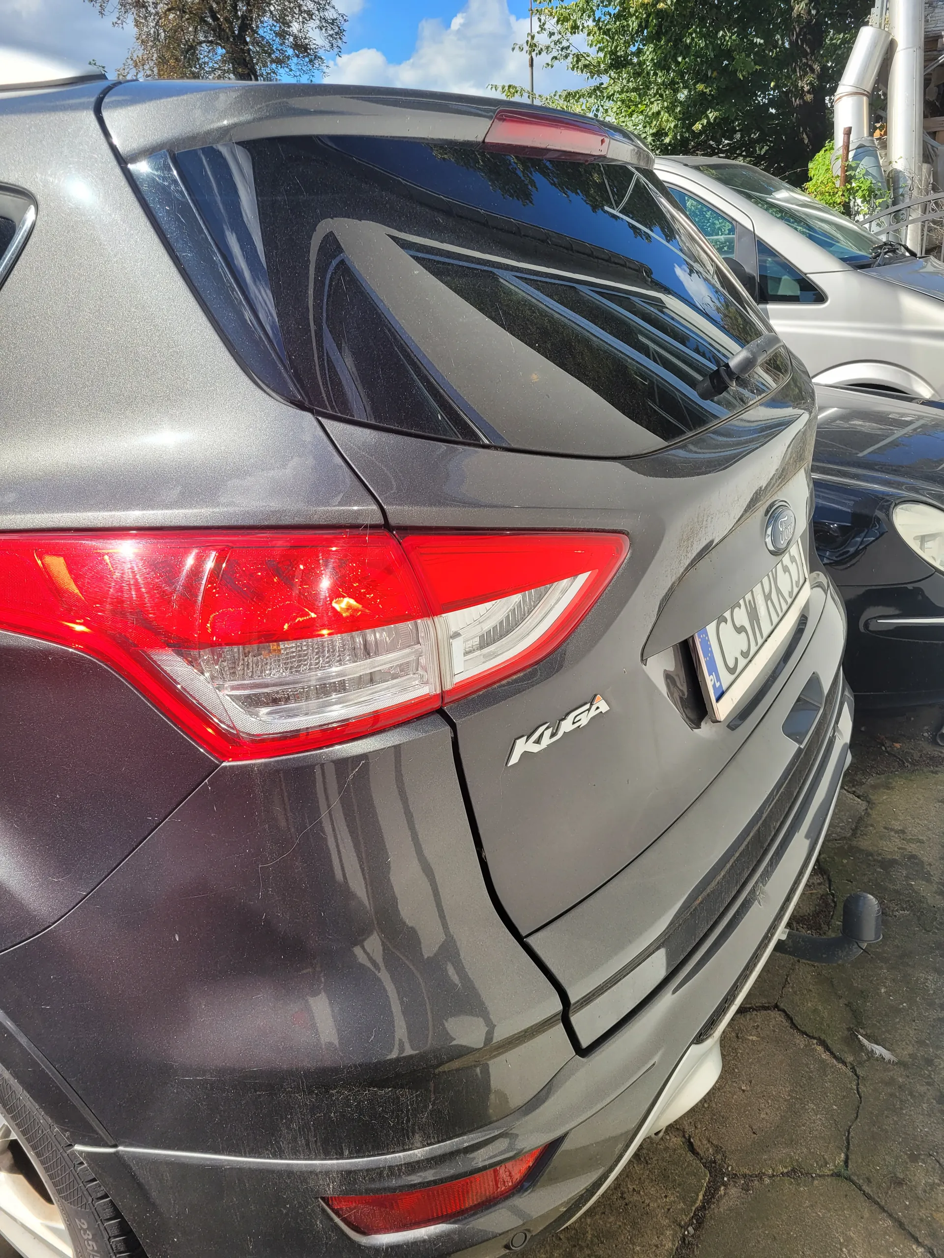 Zdjęcie przedstawia tył ciemnoszarego samochodu marki Ford model Kuga. Widoczne są czerwone światła tylnego, tablica rejestracyjna z napisem CSH 133, a także napis Kuga na bagażniku. W tle znajdują się inne zaparkowane pojazdy (srebrny i czarny), drzewa, niebo z chmurami oraz pęknięta nawierzchnia asfaltowa.