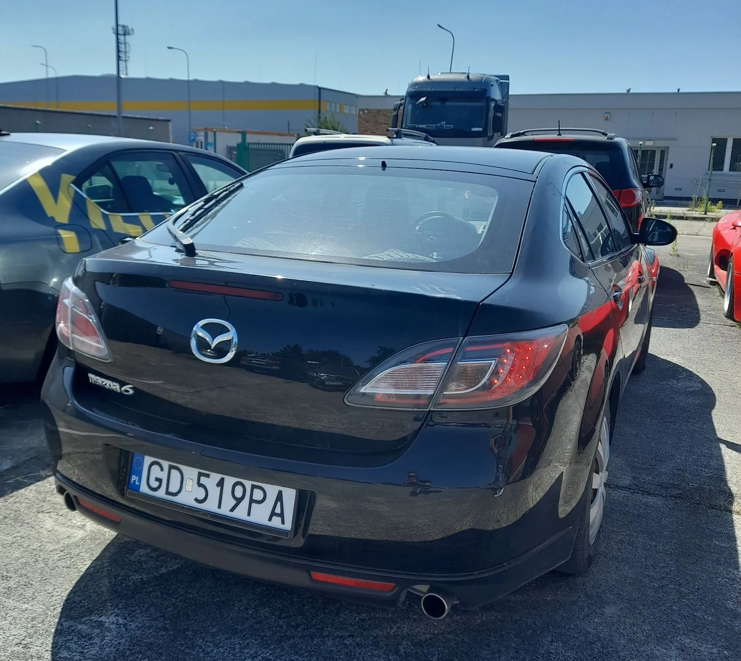 Tylny widok czarnego samochodu marki Mazda 6 zaparkowanego na placu. Na klapie bagażnika widoczne jest logo Mazdy oraz napis 'MAZDA 6'. Samochód posiada polską tablicę rejestracyjną o numerze GD 519PA. W tle widoczne są inne zaparkowane pojazdy, w tym czerwony samochód oraz ciemna ciężarówka, a także przemysłowe budynki pod jasnym, bezchmurnym niebem.