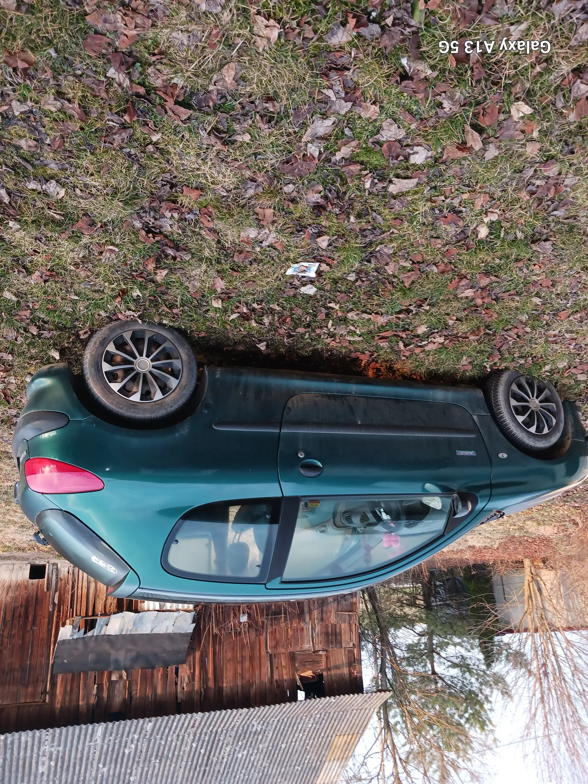 Zielony samochód osobowy zaparkowany na trawie pokrytej suchymi liśćmi. W tle drewniana budowla z blachą falistą. Widoczny tekst "Galaxy A13 5G" po prawej stronie obrazu. Wewnątrz samochodu znajdują się przedmioty. Na trawie leżą rozrzucone liście i mały biały przedmiot.