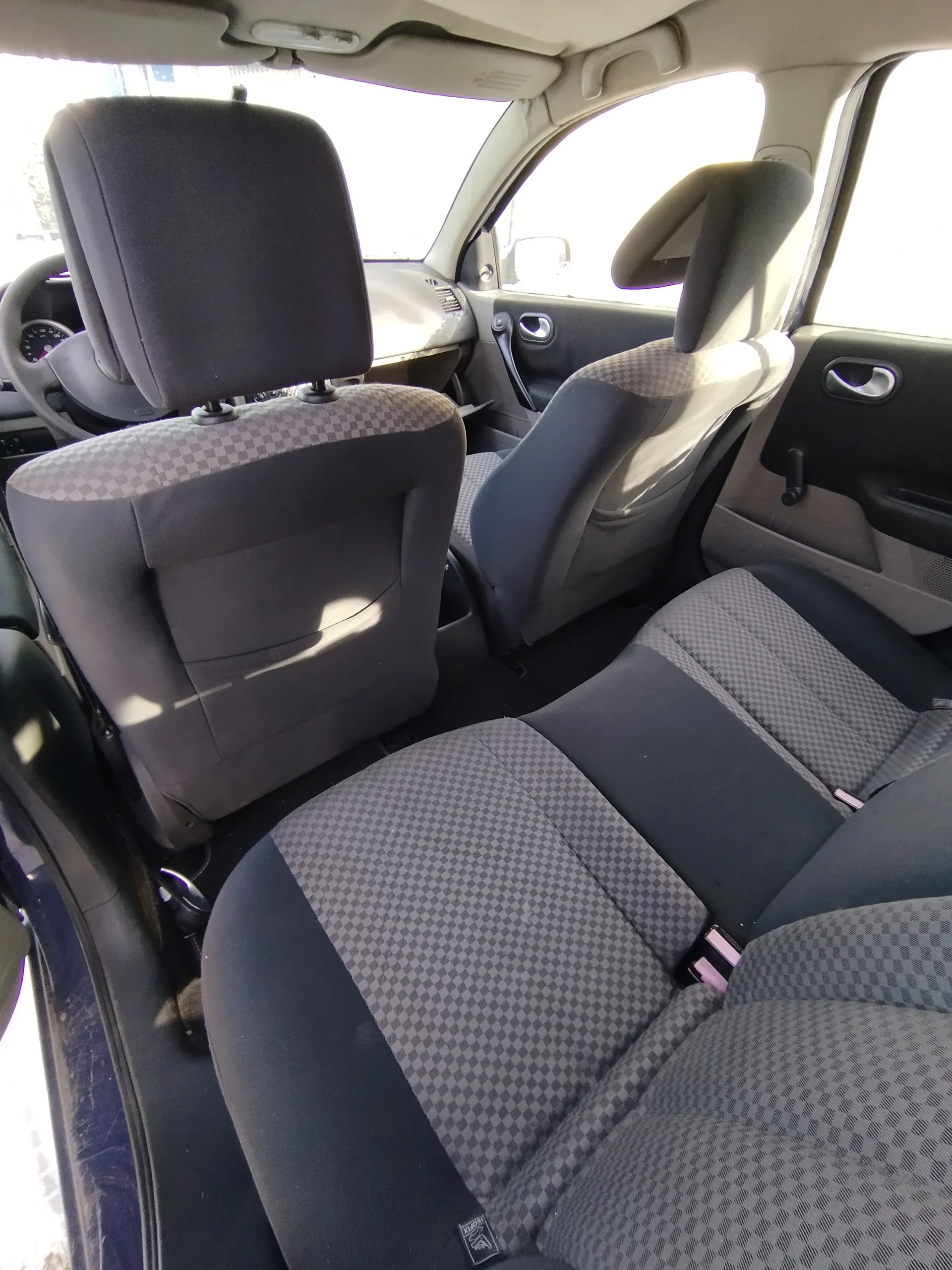 Wnętrze samochodu z widokiem na przednie siedzenia. Siedzenia pokryte tkaniną w szaro-czarną kratę. Widoczne kierownica po lewej stronie, deska rozdzielcza, drzwi z uchwytami i oknami. Wnętrze oświetlone światłem dziennym.