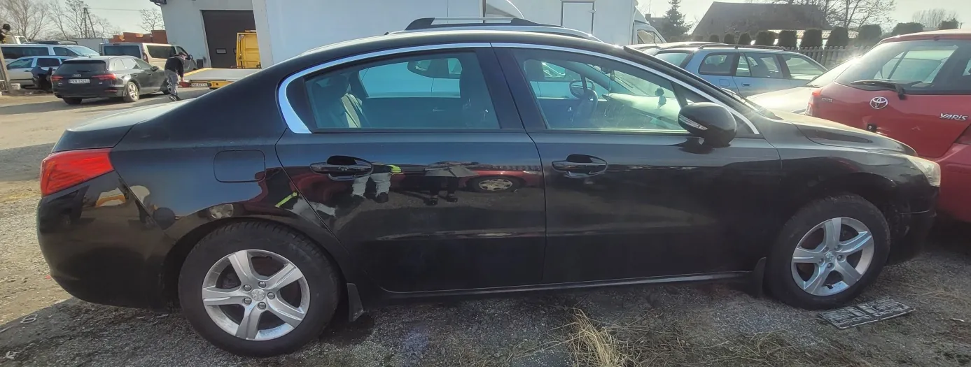 Czarny samochód osobowy typu sedan zaparkowany na otwartym placu. Po prawej stronie widoczny czerwony samochód z napisem YARIS na tylnym zderzaku. W tle inne zaparkowane pojazdy, budynek z bramą oraz osoba idąca. Na ziemi sucha trawa i nierówny asfalt. Odbicia na błyszczącej karoserii czarnego samochodu.