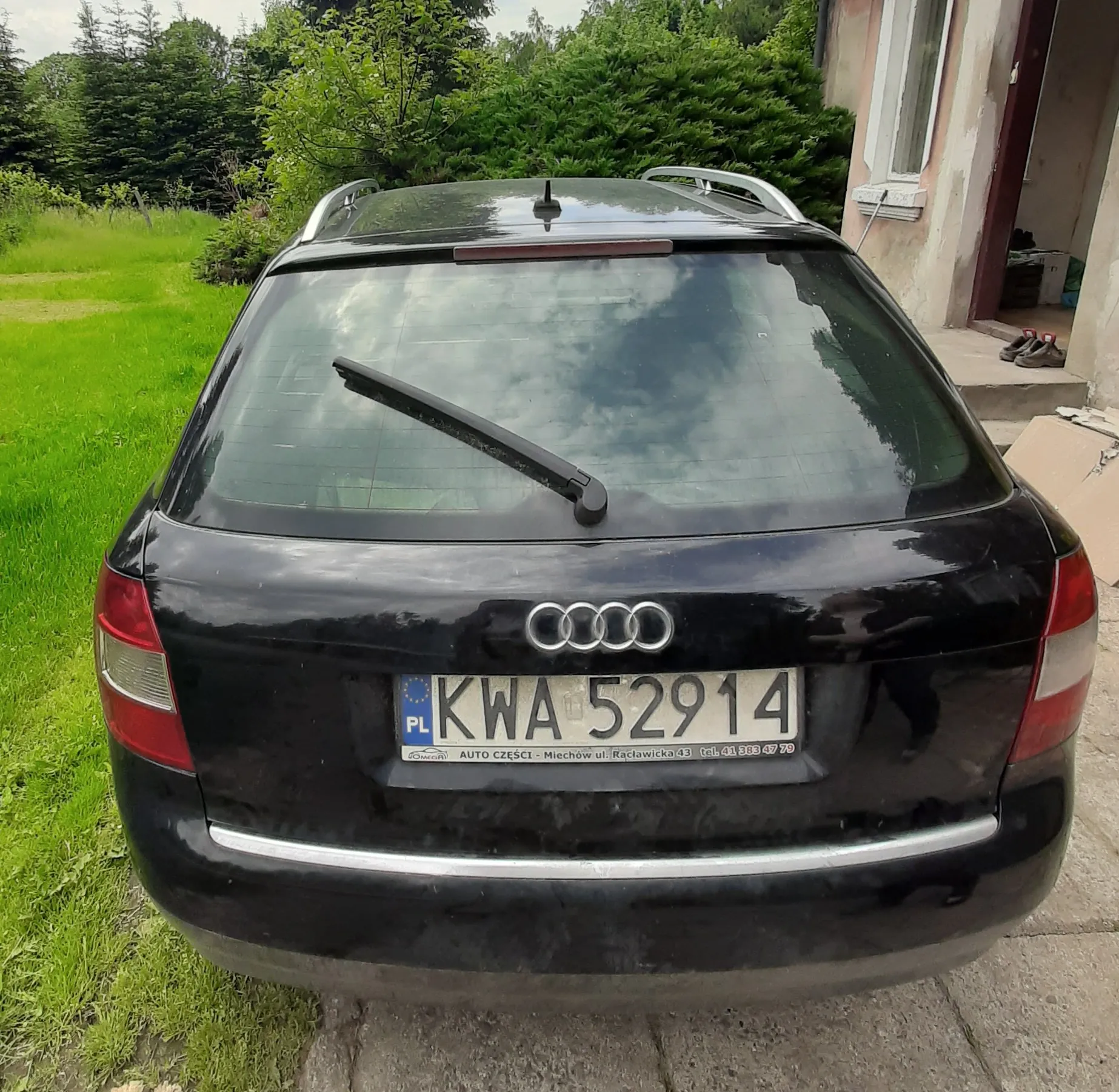 Tylny widok czarnego samochodu marki Audi w nadwoziu kombi. Pojazd posiada srebrne relingi dachowe i tablicę rejestracyjną o numerze KWA 52914 z oznaczeniem PL. Samochód zaparkowany jest na betonowym podjeździe, w tle widoczna jest zielona trawa, drzewa oraz fragment budynku mieszkalnego.