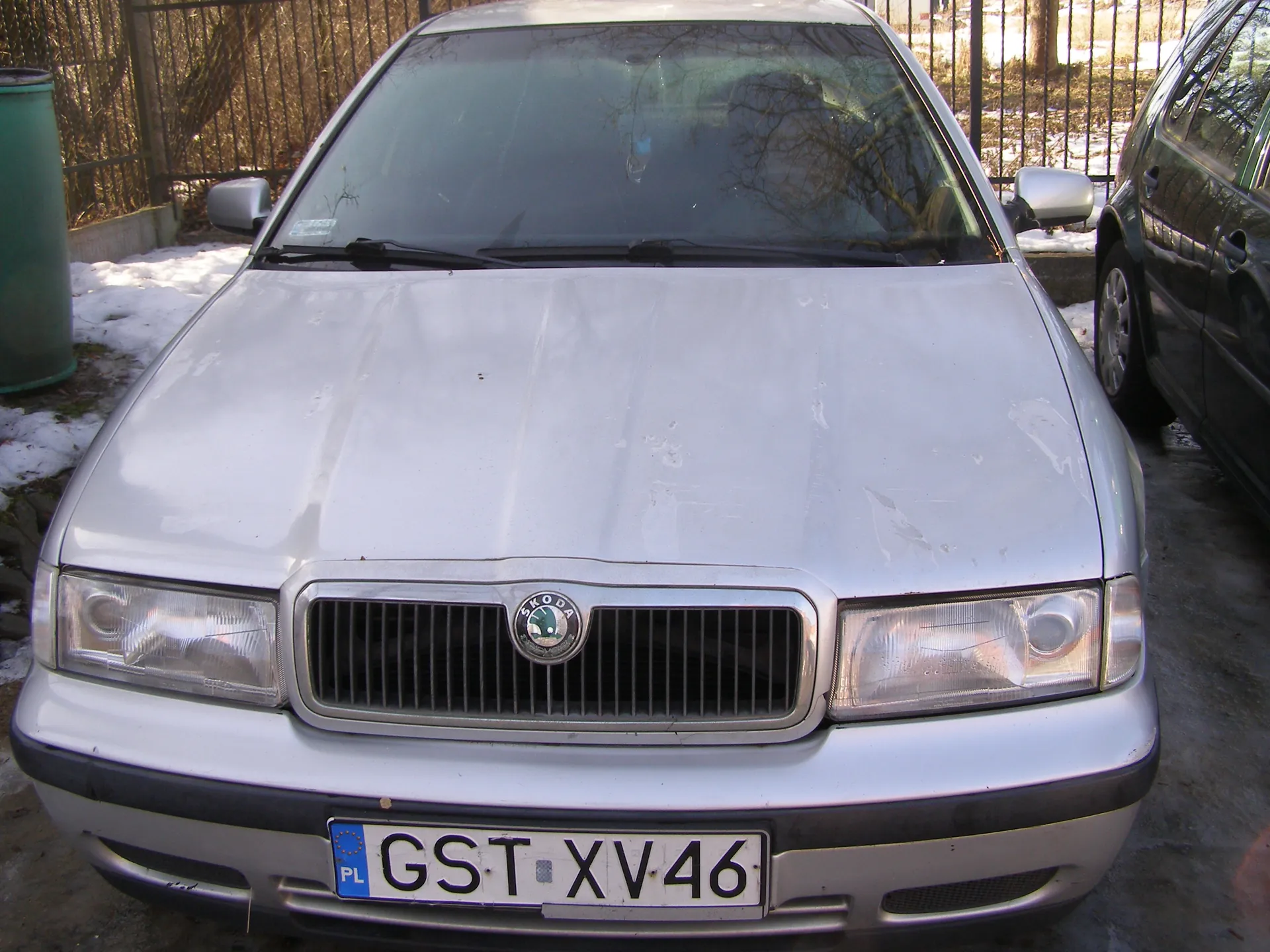 Przód srebrnego samochodu marki Škoda z tablicą rejestracyjną GST XV46 (PL) umieszczoną na środku pasa przedniego. Na masce widoczny emblemat marki. Samochód zaparkowany na zewnątrz, w tle metalowe ogrodzenie, śnieg na ziemi oraz fragment czarnego samochodu po prawej stronie. W tle również suche krzewy i drzewa.
