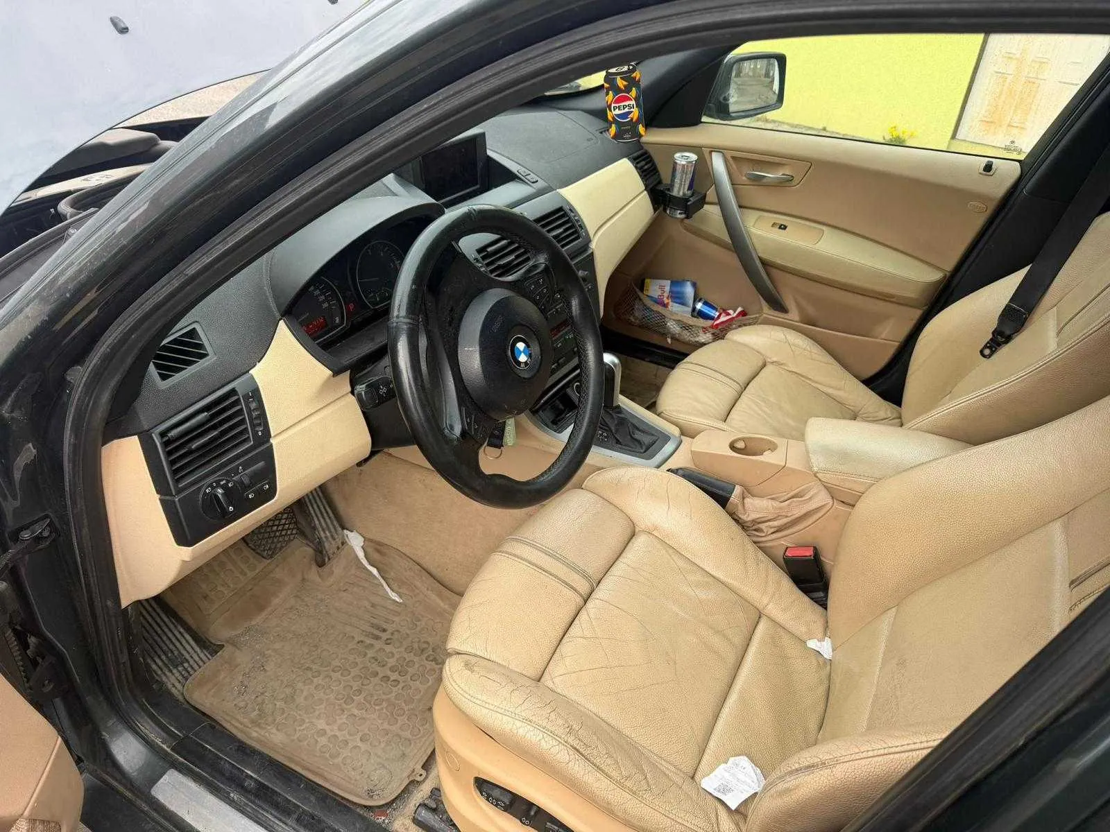 Wnętrze samochodu marki BMW z beżowymi skórzonymi fotelami i czarną kierownicą z logo BMW. Deska rozdzielcza w kolorze beżowo-czarnym, widoczne zanieczyszczone dywaniki podłogowe. Na desce rozdzielczej stoi puszka Pepsi, w uchwycie drzwiowym znajduje się puszka Red Bull. Na siedzeniu pasażera leżą różne przedmioty, w tym plastikowa torba i butelka. Drzwi kierowcy są otwarte, na zewnątrz widoczny żółty budynek i fragment białej konstrukcji. W samochodzie znajdują się również białe ręczniki lub papierowe kawałki na siedzeniach i podłodze.