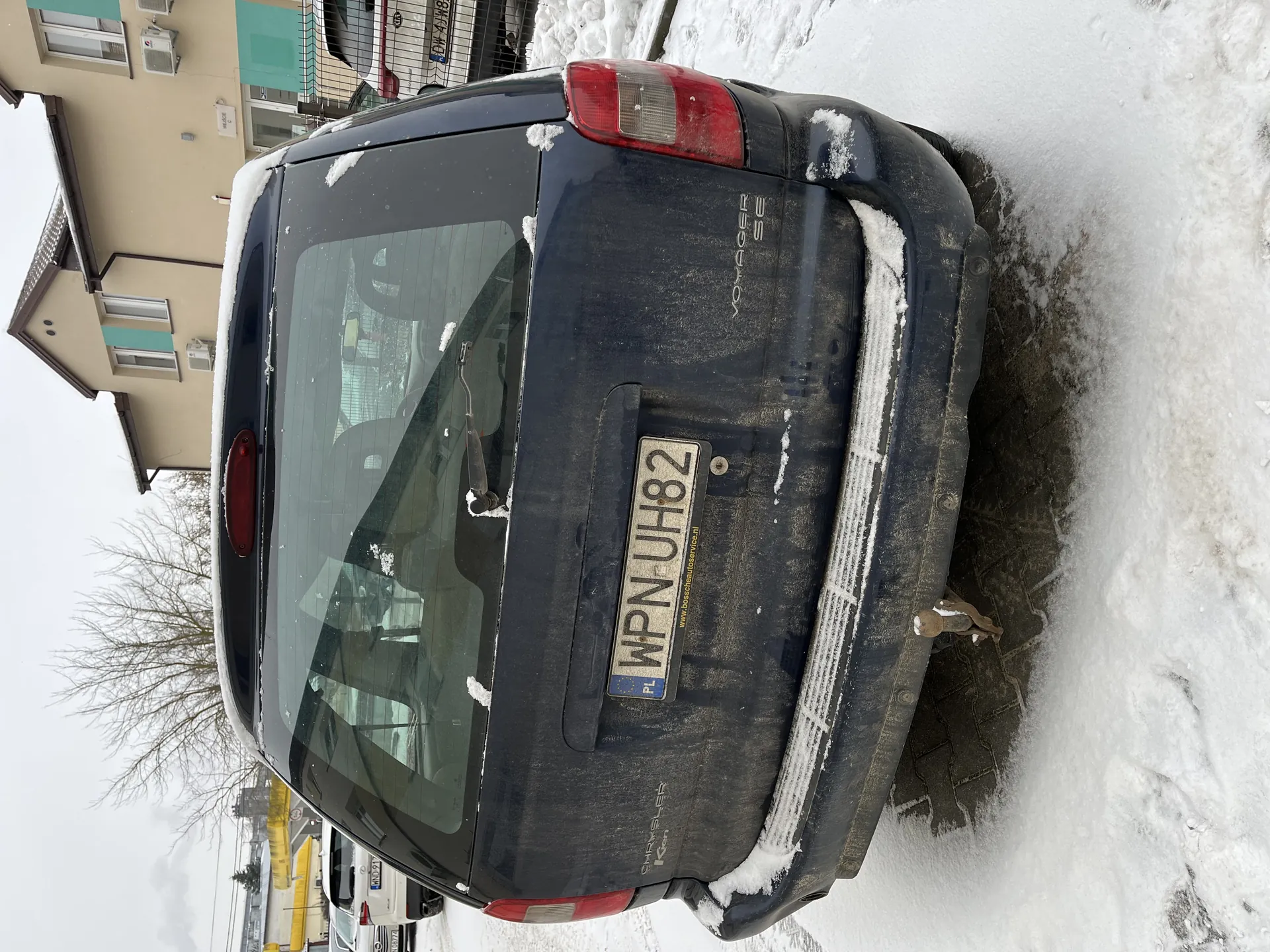 Z tyłu ciemny samochód Voyager SE z tablicą rejestracyjną WPN UH82 (PL), pokryty śniegiem na szybie i bagażniku. W tle budynki mieszkalne z klimatyzatorami, drzewo bez liści i śnieżna droga. Widoczne są inne pojazdy oraz siatka ogrodzeniowa.