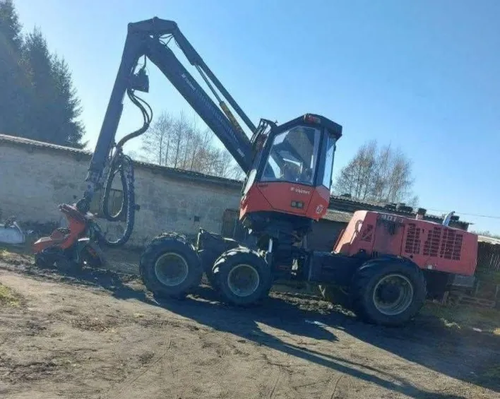 Czerwona maszyna leśna z dużym, hydraulicznym ramieniem i chwytakiem, osadzona na czterech dużych kołach terenowych. Maszyna znajduje się na piaszczystym podłożu na tle niskiego muru i drzew pod jasnym, niebieskim niebem.