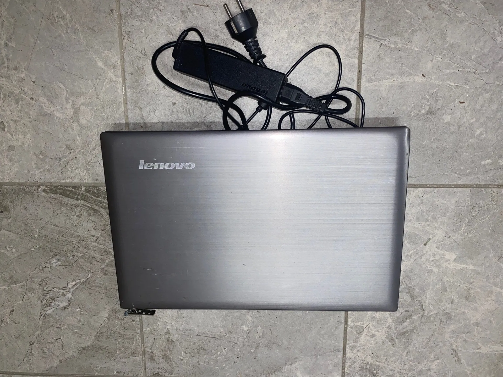 Srebrny laptop marki Lenovo zamknięty i leżący na szarych płytkach ceramicznych. Nad laptopem znajduje się czarny zasilacz z kablem i wtyczką sieciową.