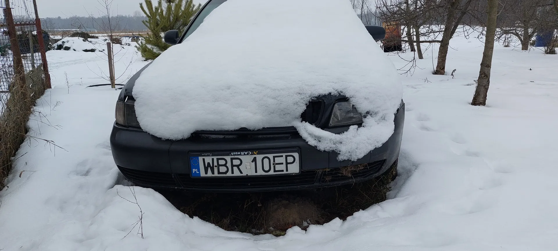 Czarny samochód pokryty grubą warstwą śniegu, widok od przodu. Na tablicy rejestracyjnej widoczny napis WBR 10EP oraz oznaczenie PL po lewej stronie. W tle śnieżna sceneria z drzewami bez liści, ogrodzeniem i śladami po oponach na śniegu. Niebo jest zachmurzone.