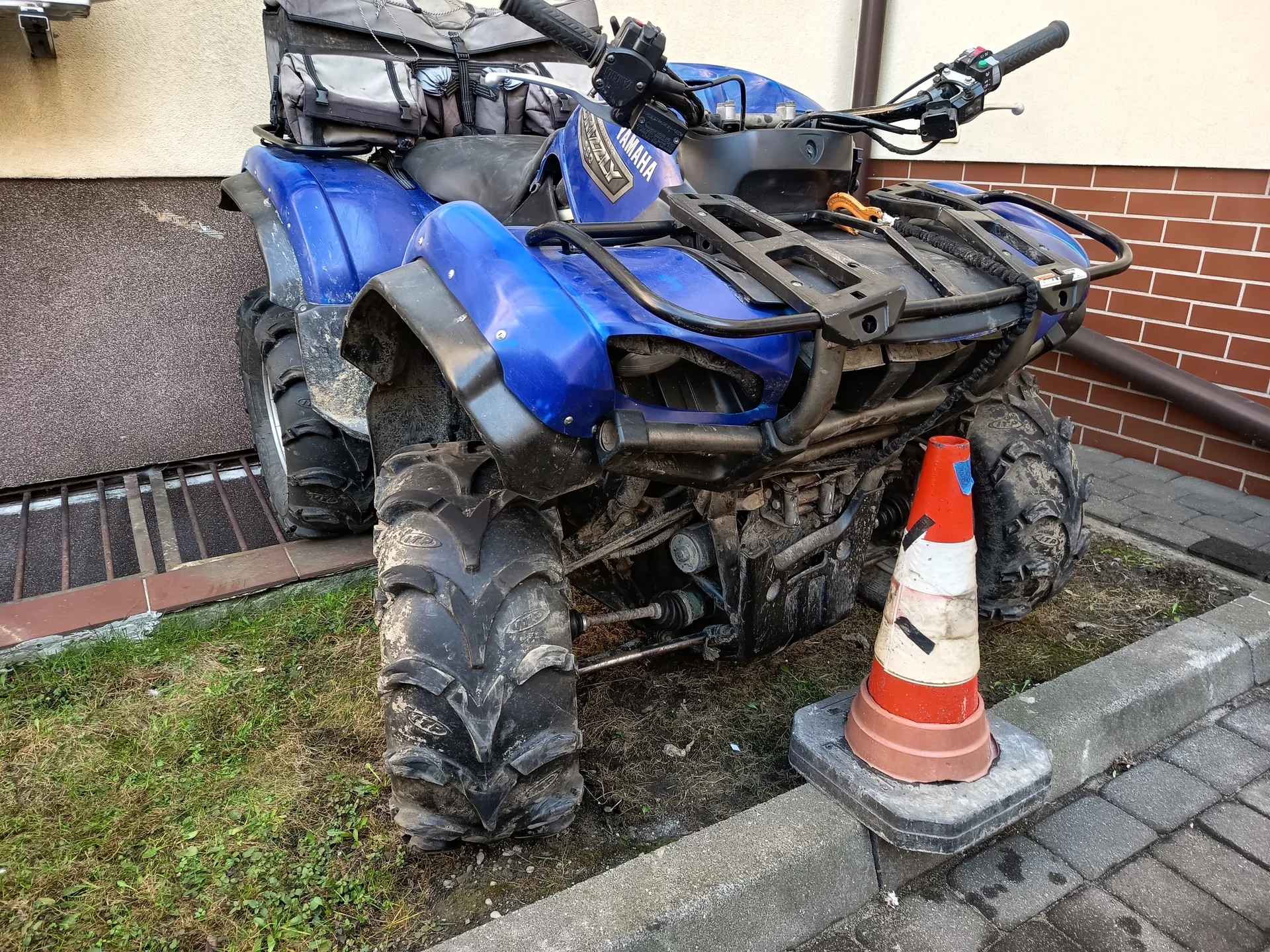 Niebieski quad marki YAMAHA z brudnymi oponami, zaparkowany przy budynku z ceglaną ścianą i beżowym tynkiem. Obok quadu stoi pomarańczowo-biały stożek drogowy z czarnymi znakami, umieszczony na betonowej podstawie. W tle widoczna trawa, brukowana ścieżka oraz metalowa kratka odpływowa. Na quadzie zamocowane są torby, a na jego nadwoziu widnieje napis YAMAHA.