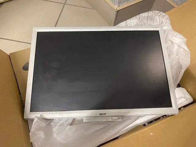 Monitor Acer o srebrnej ramie z czarnym ekranem umieszczony w kartonowym pudełku na białej folii. W tle widoczna podłoga z kafelkami. Logo 'acer' znajduje się po lewej stronie ramy monitora.